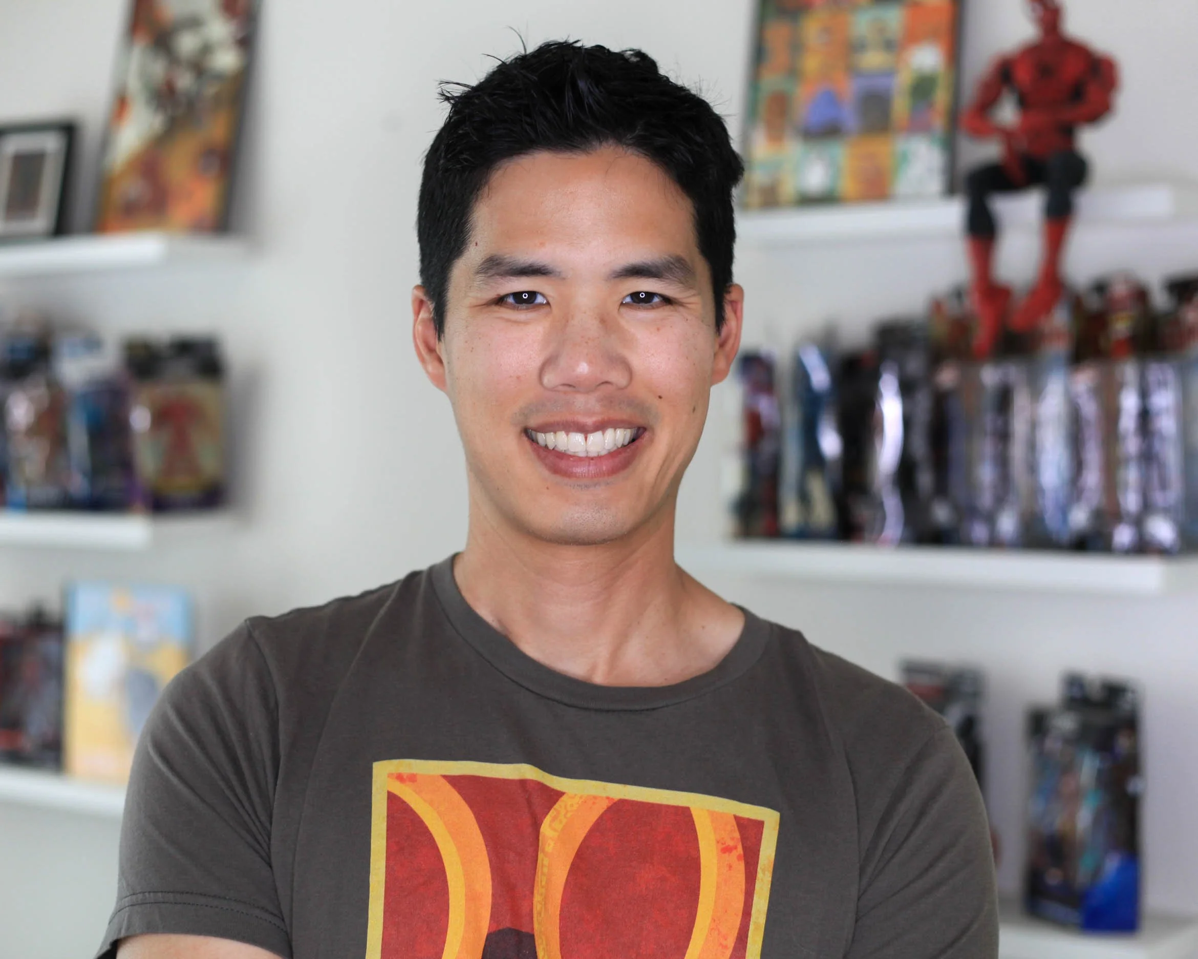 A young man with short black hair and a wide smile is wearing a dark superhero-themed T-shirt. The background shows bookshelves with colorful books and a Batman figurine.