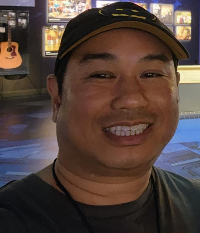A smiling man wearing a black baseball cap with yellow accents, in a room with multiple TV screens and guitars in the background.