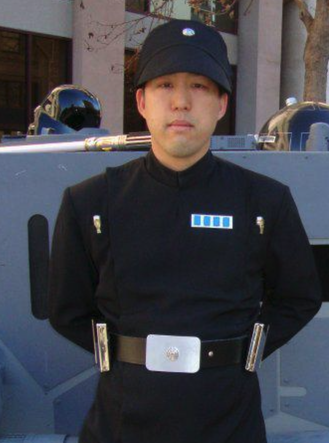 Person dressed as Darth Vader from Star Wars standing outdoors in front of a vehicle, wearing a black uniform and hat.