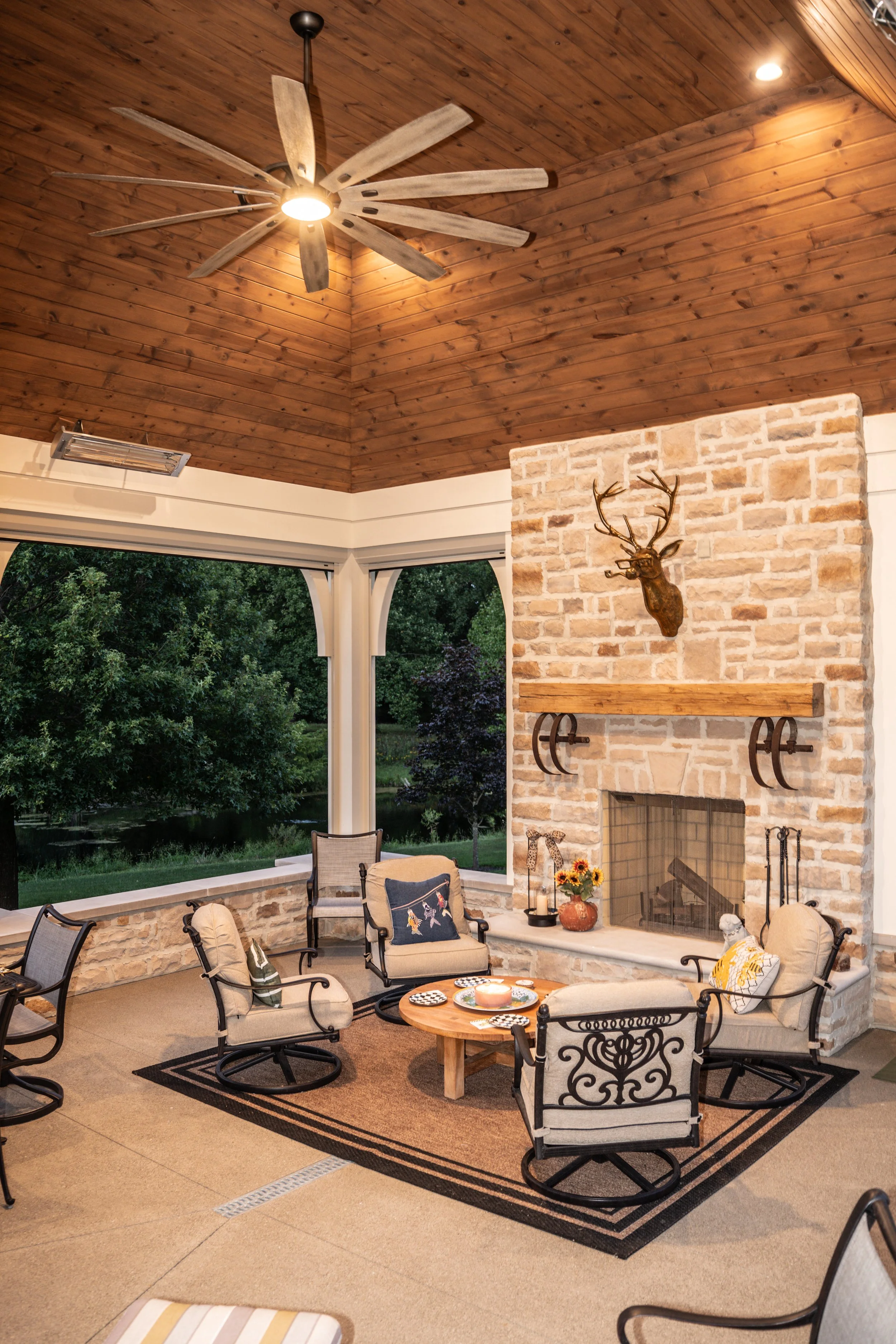 A cozy outdoor living room with beige cushioned chairs, a small round wooden table, and a black patterned rug. There is a brick fireplace with a deer head mounted above it. A ceiling fan hangs from a wooden ceiling, and large open windows reveal lush greenery outside.