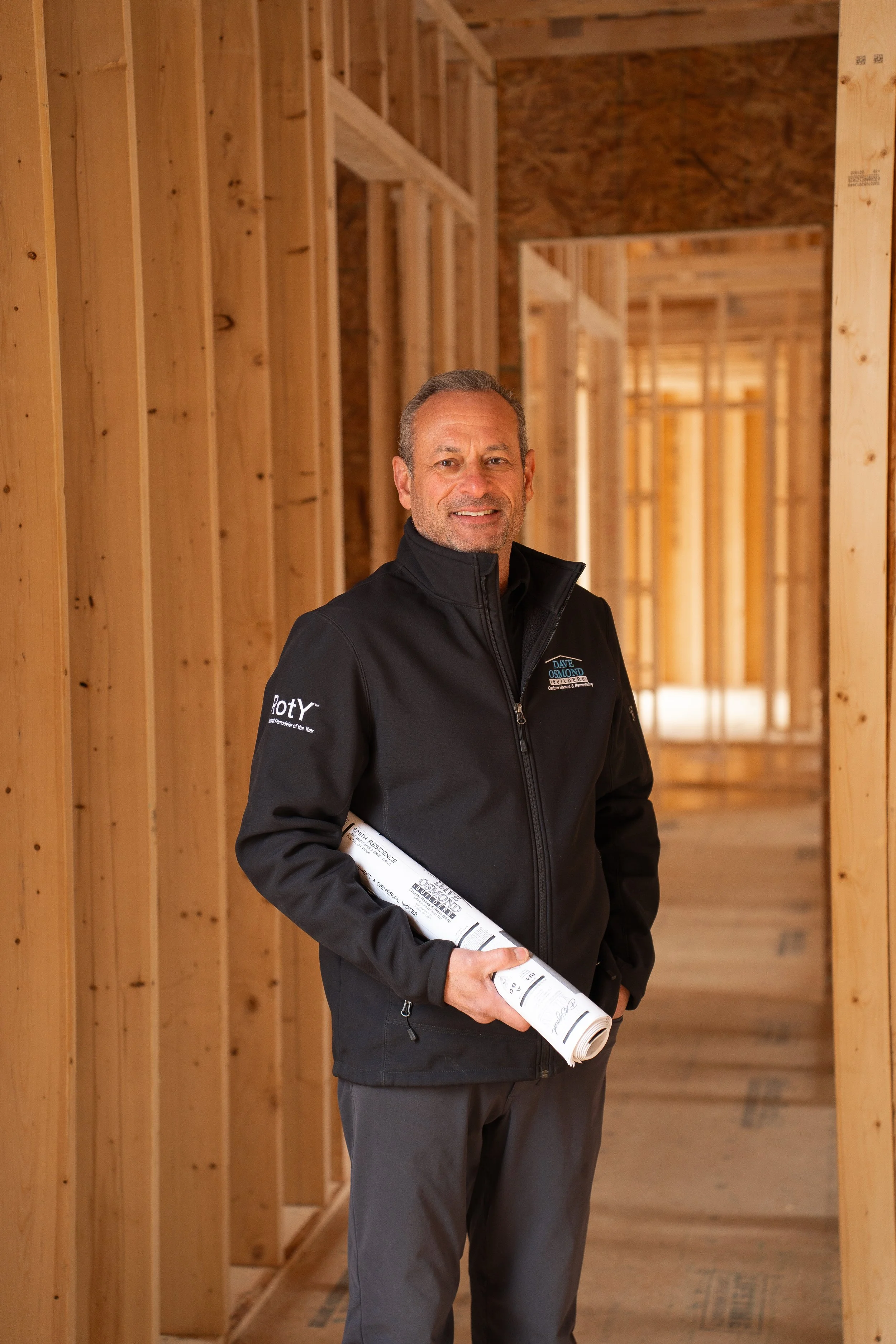 A man in a black jacket is standing inside a wooden building under construction, holding rolled-up blueprints and smiling.