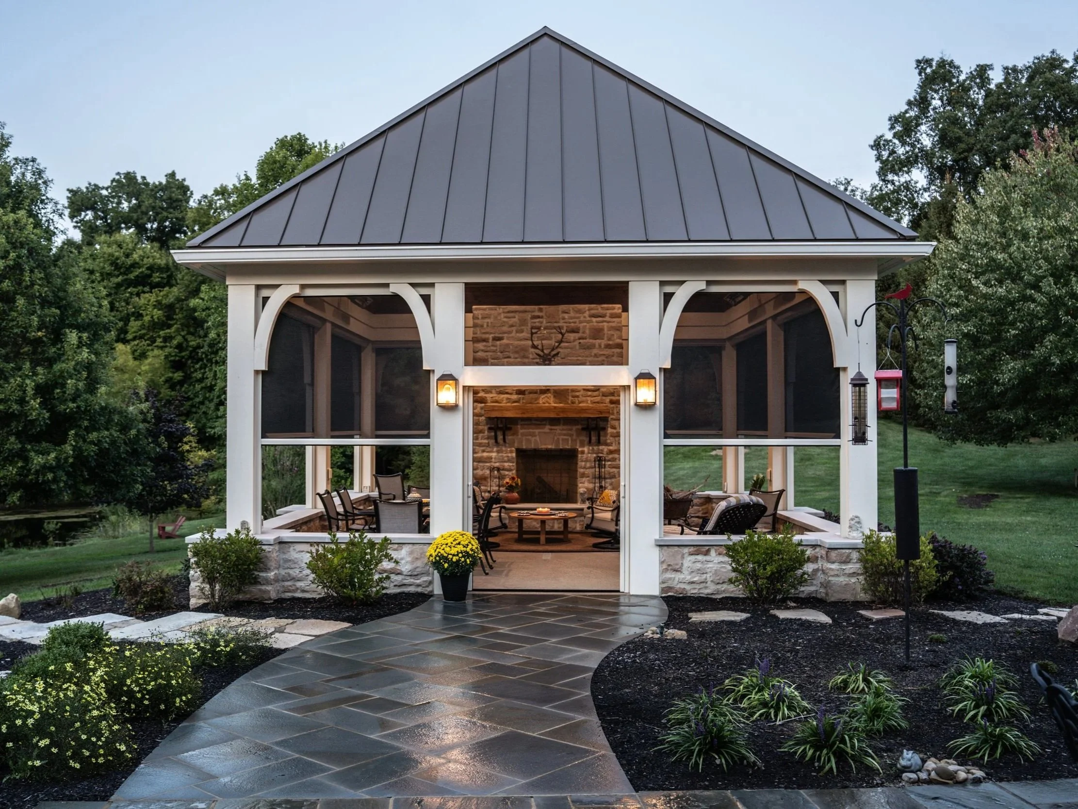 A backyard patio with a screened gazebo, stone fireplace, outdoor seating, potted plants, garden beds, and garden lighting in the evening.