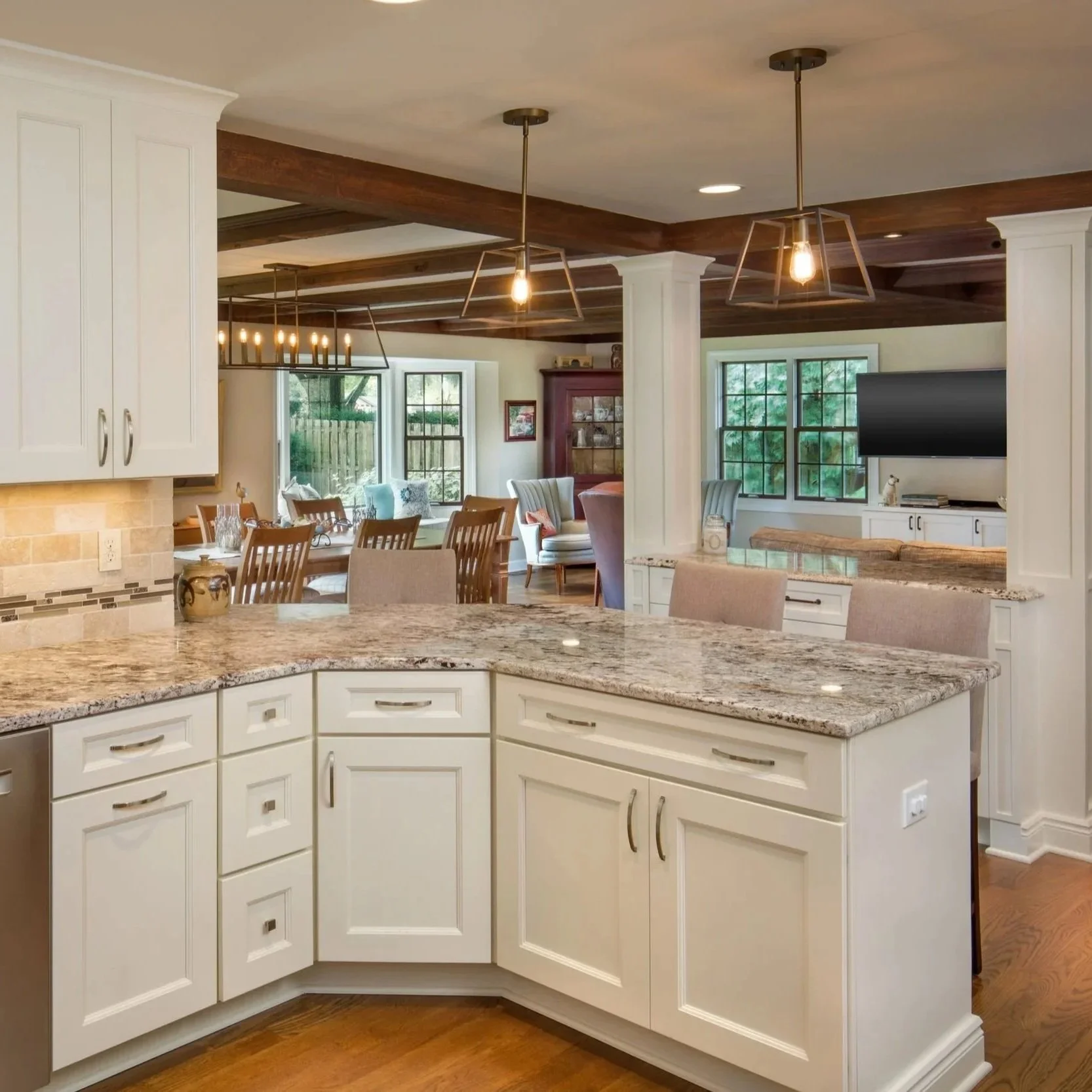 Kitchen with white cabinets and granite countertops, wooden dining table with chairs, large windows, and a living area with a TV and seating in the background.