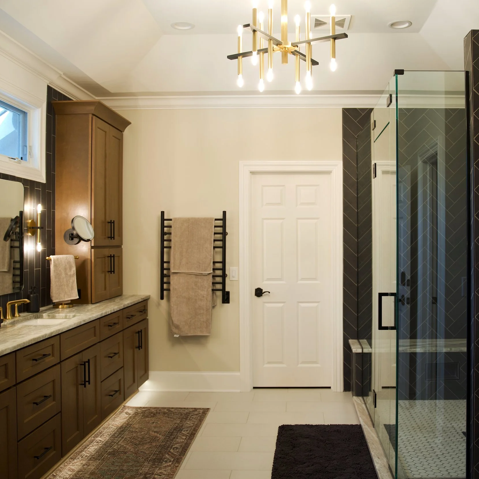 Modern bathroom with a walk-in glass shower, black tile accents, white hexagon tile floor, beige walls, and a wooden cabinet with a marble countertop. Two beige towels hang on a black towel rack, and a black rug is on the floor. A ceiling light fixture with multiple bulbs illuminates the space.