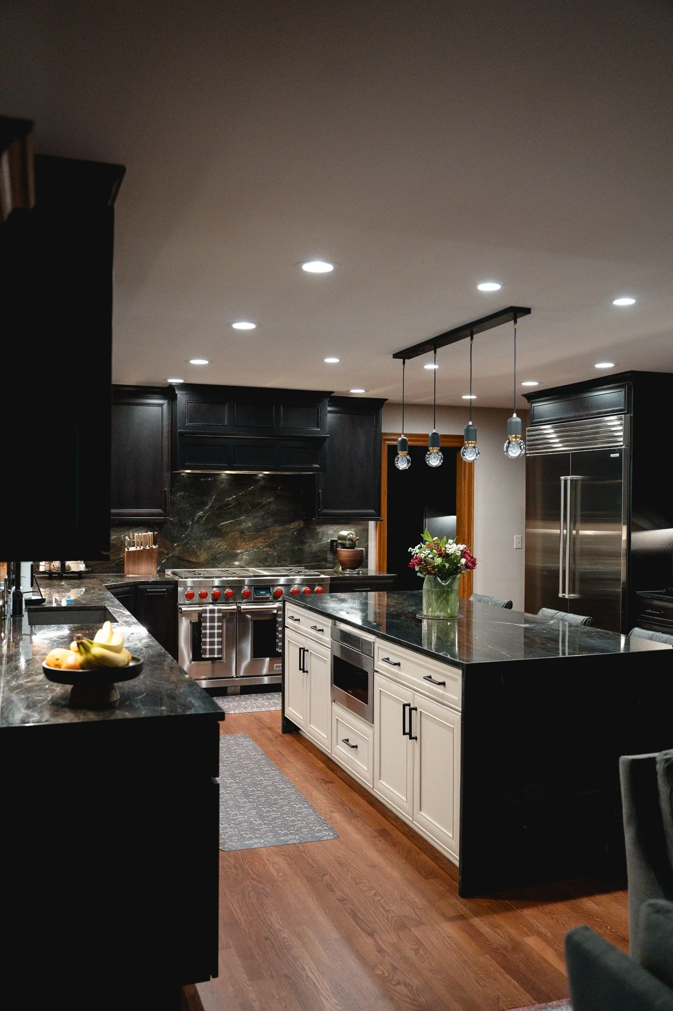 A modern kitchen with black upper cabinets, white lower cabinets, a black countertop, a stainless steel oven, a refrigerator, and a pendant light fixture with four hanging lights above the kitchen island. There is a bowl with bananas on the counter and a vase with flowers on the island. The flooring is wood and there is a gray rug on the floor.