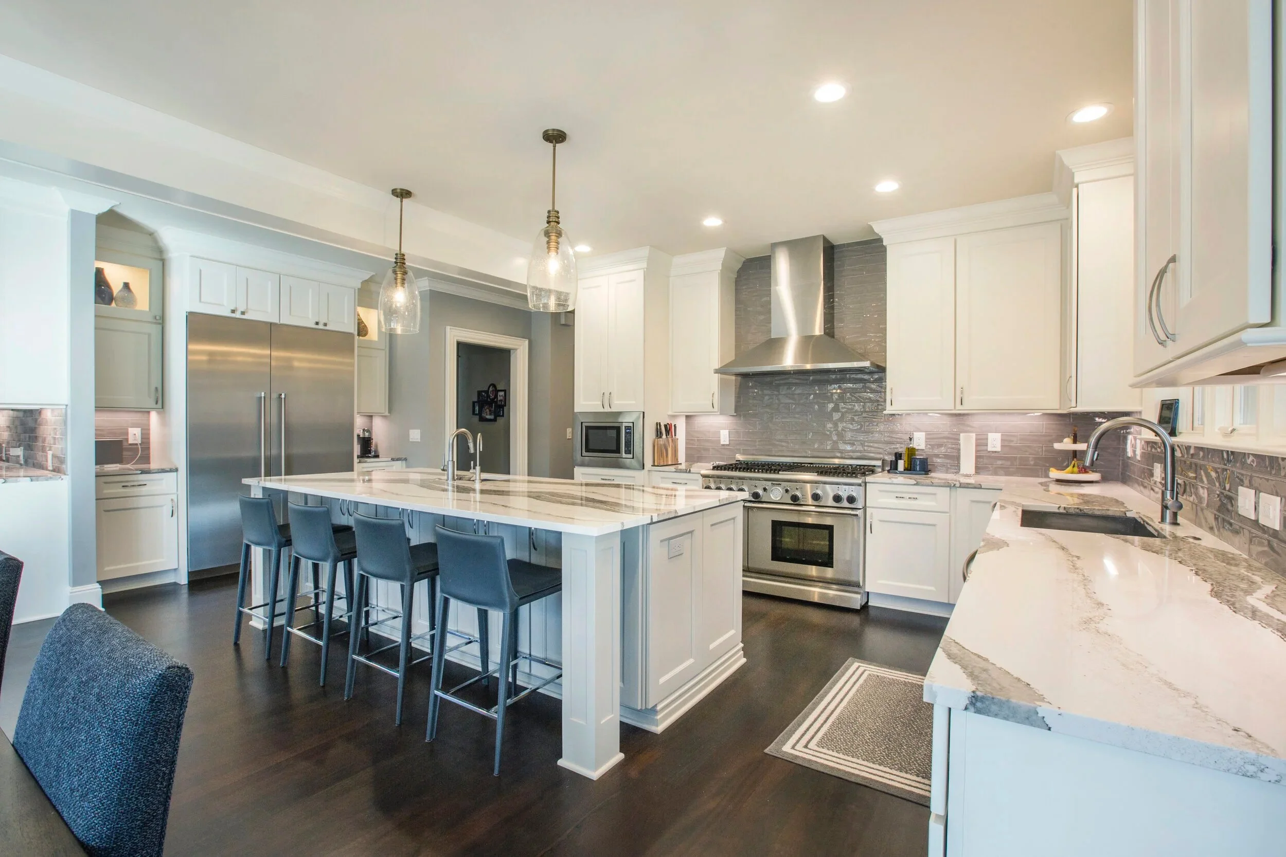 Modern white kitchen with a large island, stainless steel appliances including a refrigerator and oven, gray brick backsplash, pendant lights, and dark hardwood floors.