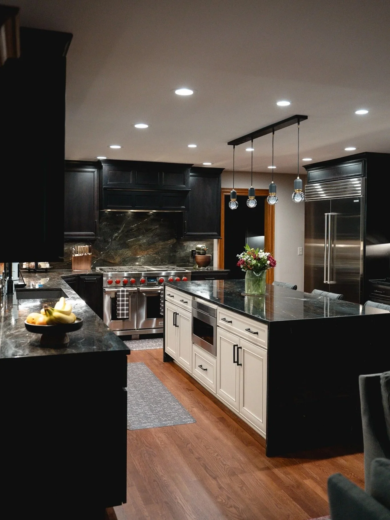 Modern kitchen with black cabinets, white island with black countertop, stainless steel appliances, a vase of flowers, and a fruit bowl on the counter.