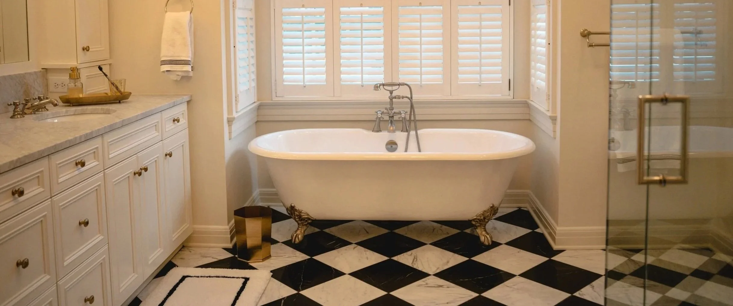Bathroom with a clawfoot bathtub in front of a window with white shutters, white cabinets on the left, patterned black and white floor, glass shower door on the right.