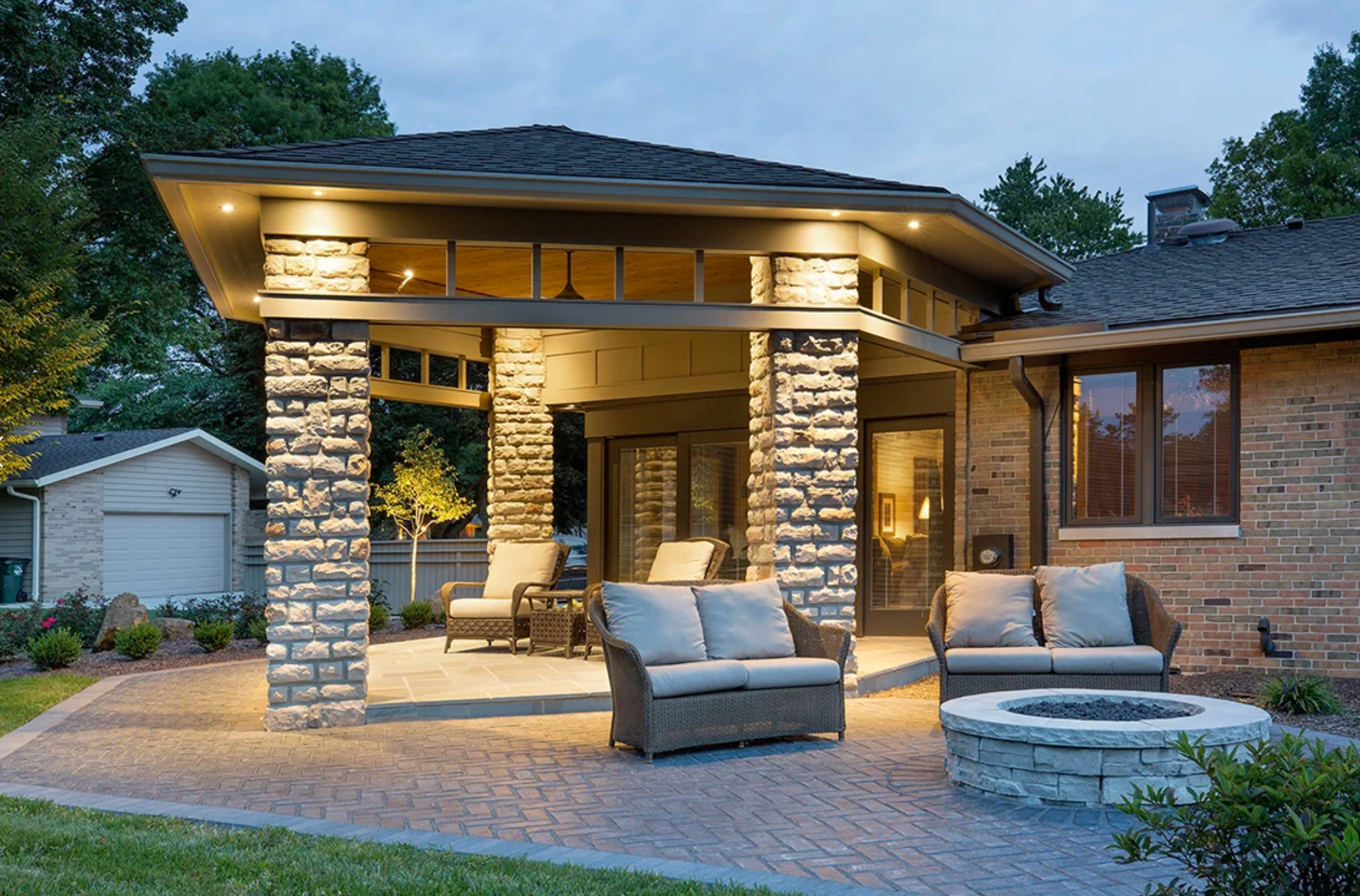 View of a backyard patio with outdoor seating, brick fire pit, and house with illuminated windows and accent lighting under a covered porch at dusk.