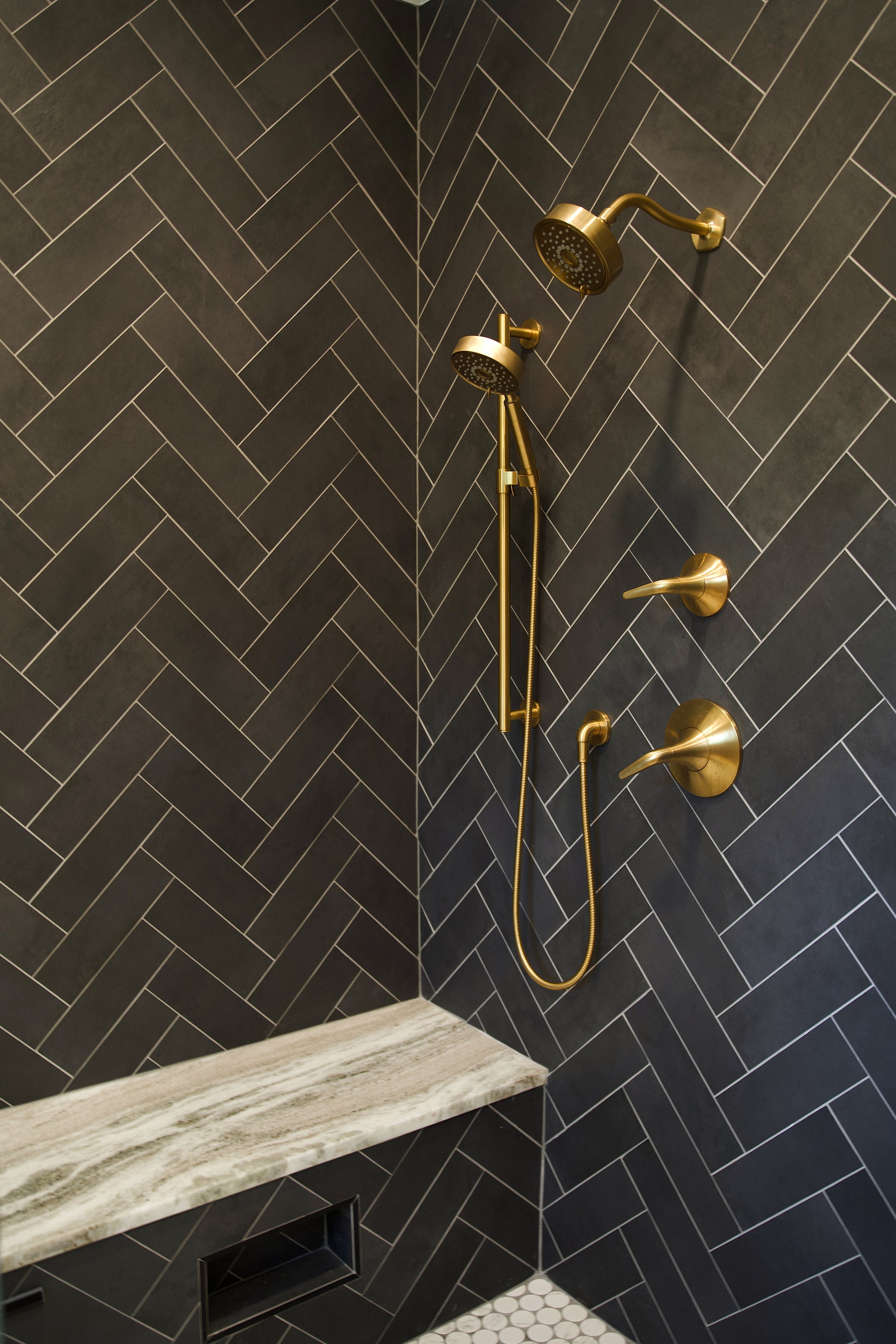 A shower area with black herringbone tiles on the walls, a brass showerhead, and a shower handle. There is a marble shelf and white pebble flooring in the corner.
