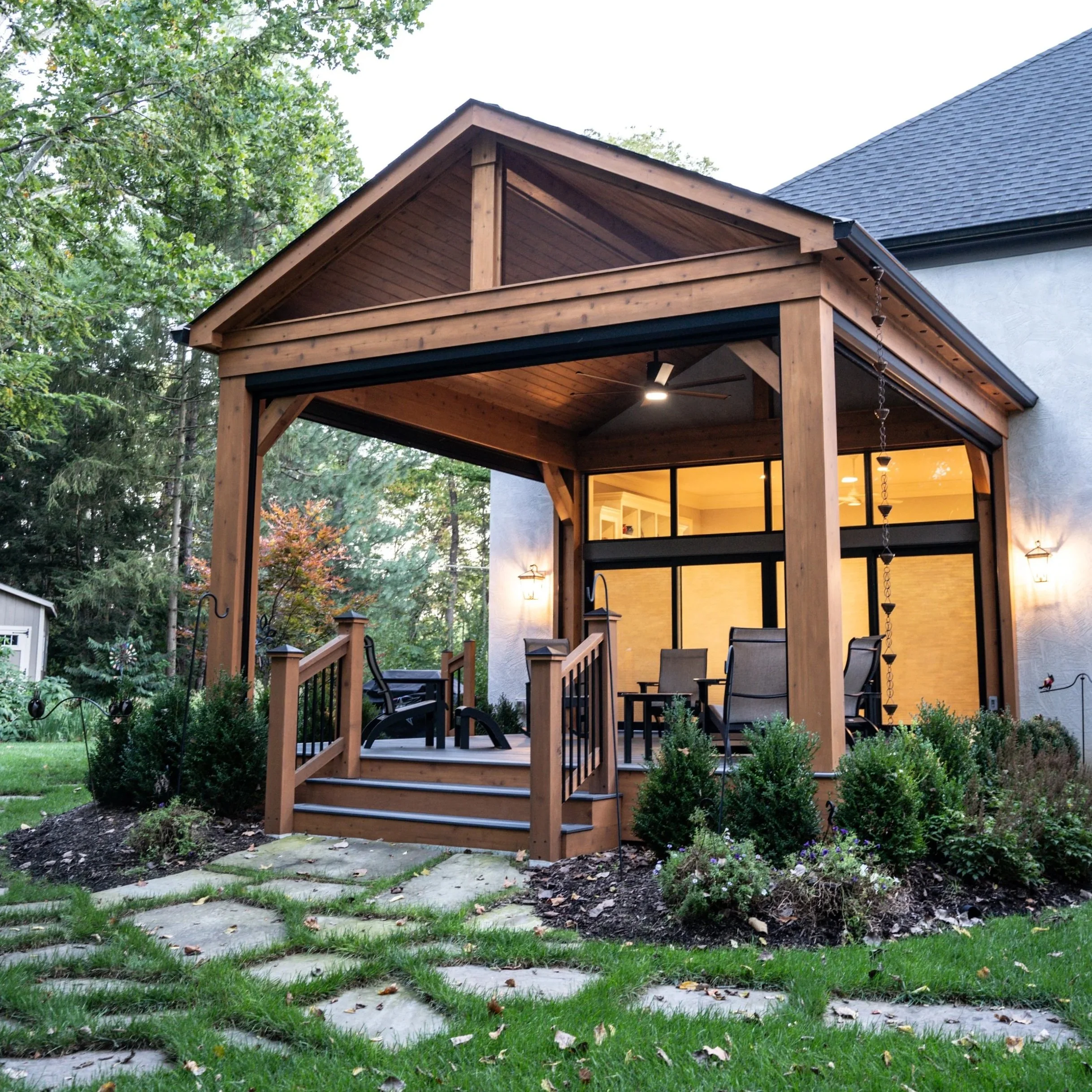 A backyard with a wooden covered patio area, surrounded by trees and greenery, with a view of a modern house.