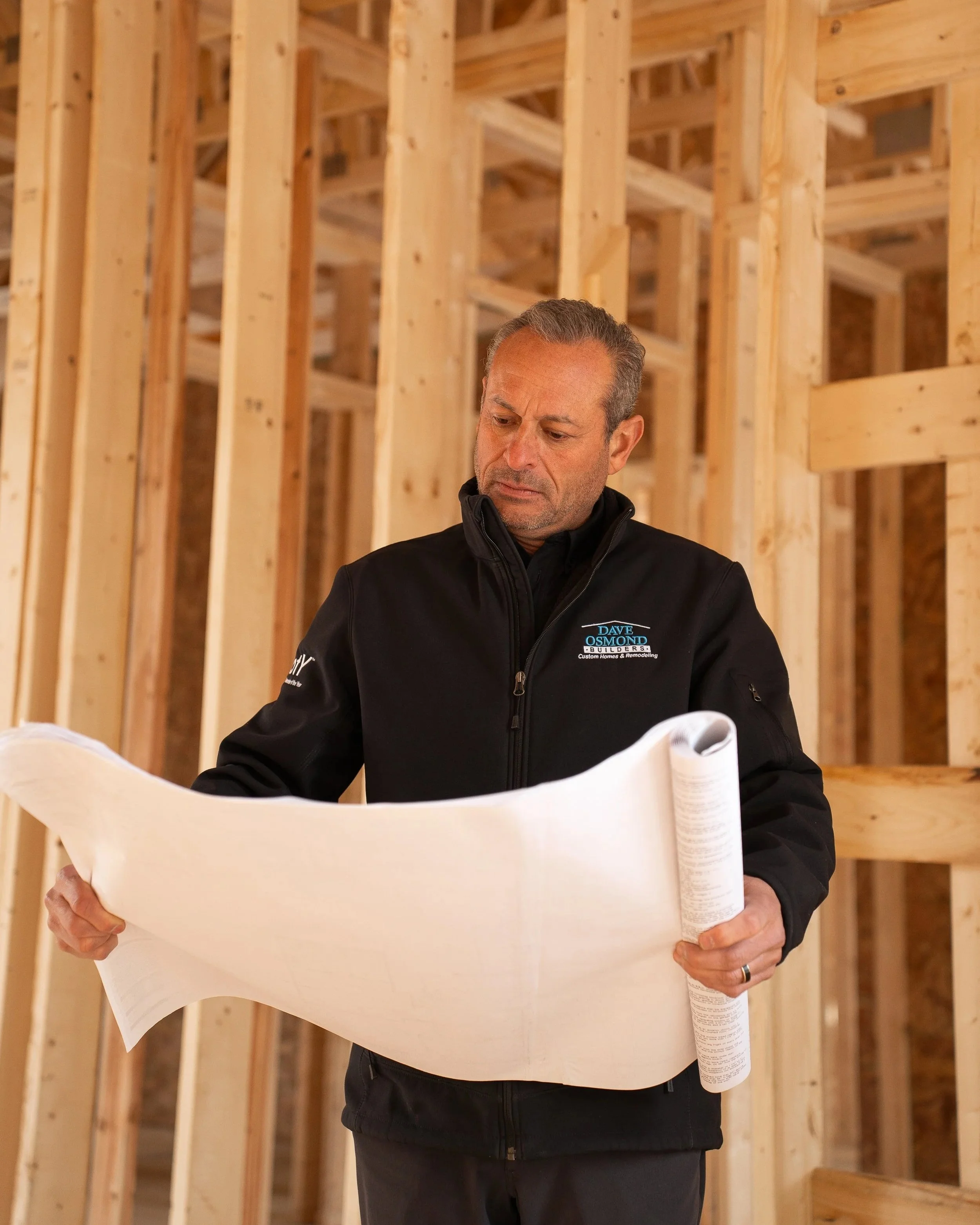 A man in a black jacket examining blueprints inside a wooden frame of a building under construction.