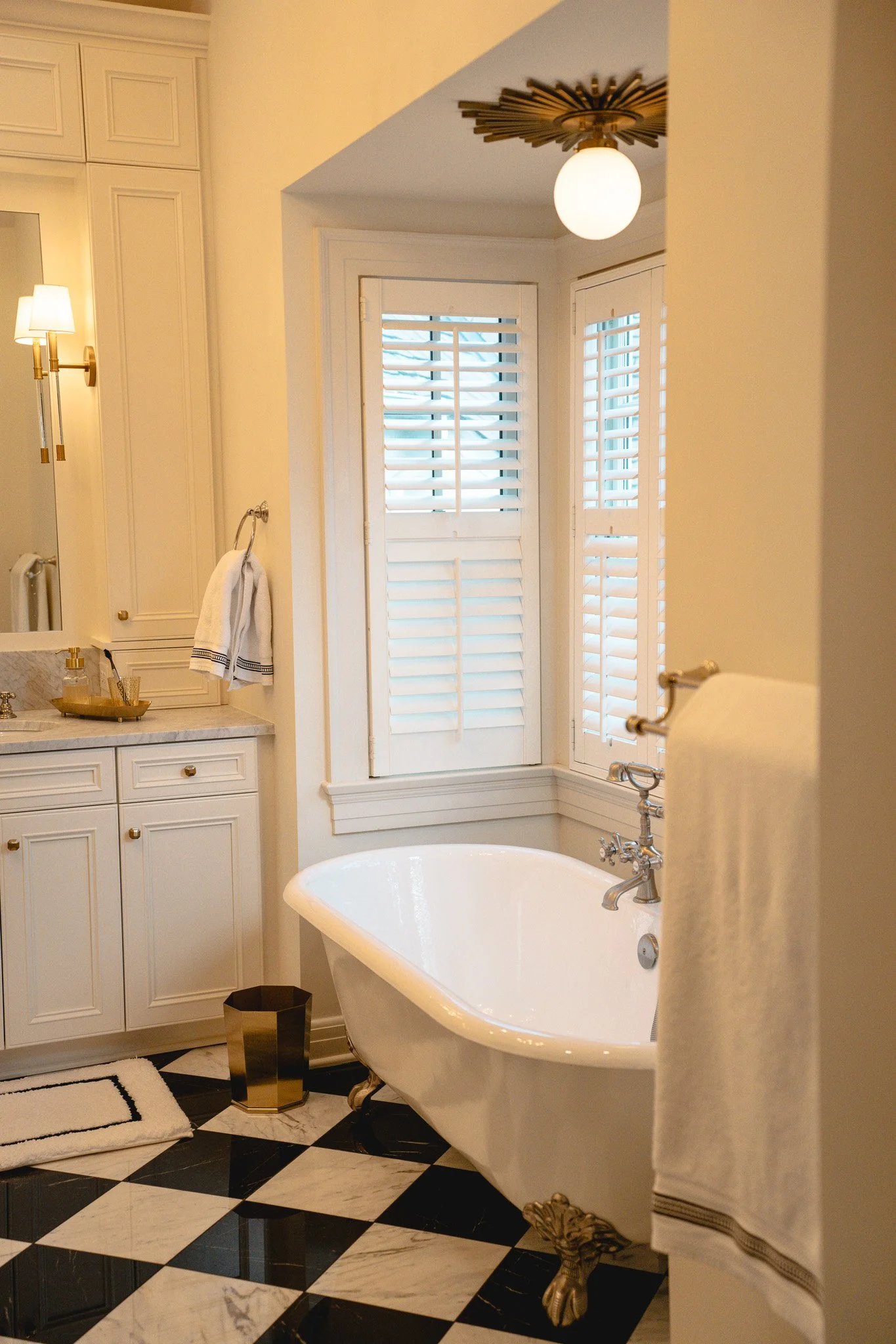 Bathroom with a clawfoot bathtub, black and white checkered floor, white cabinetry, gold fixtures, and a window with plantation shutters, illuminated by a globe ceiling light and wall sconce.