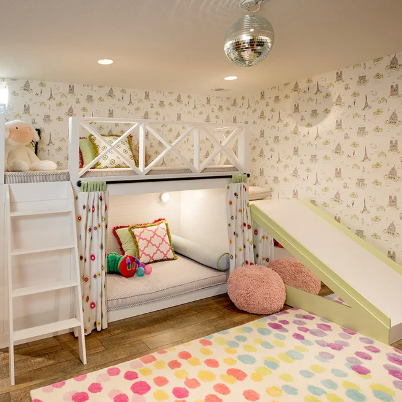 Children's playroom with a white bunk bed, climbing ladder, window curtains with colorful dots, pink fuzzy cushions, plush toys, and a polka-dot rug. The wallpaper has a Paris-themed pattern with the Eiffel Tower.