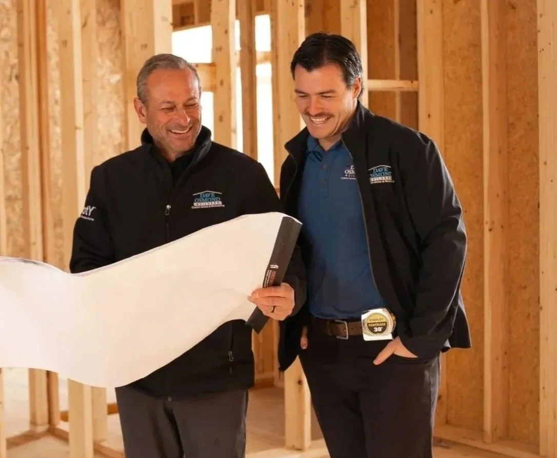 Two men looking at a blueprint inside a wooden framing of a house, both smiling and wearing black jackets with company logos.