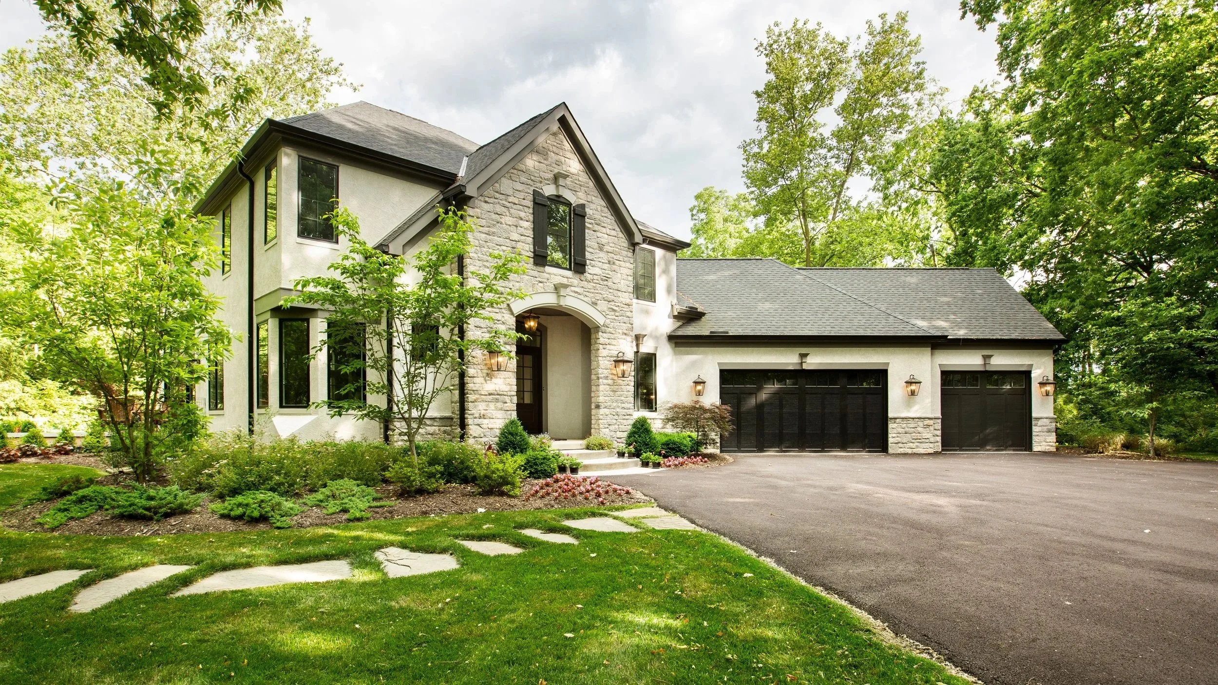 A modern two-story house with a black garage door, stone and stucco exterior, surrounded by trees, shrubs, and a landscaped yard, with a paved driveway in front.