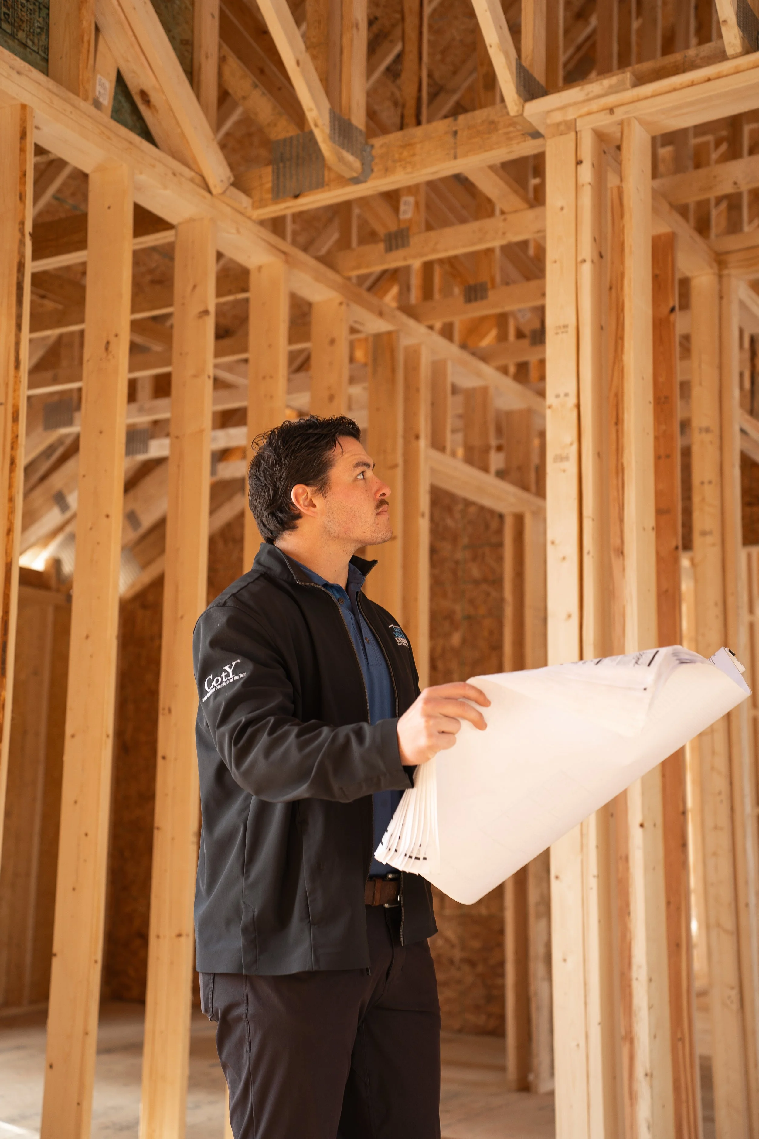 A man in a black jacket inspecting plans at a construction site with exposed wooden framing.