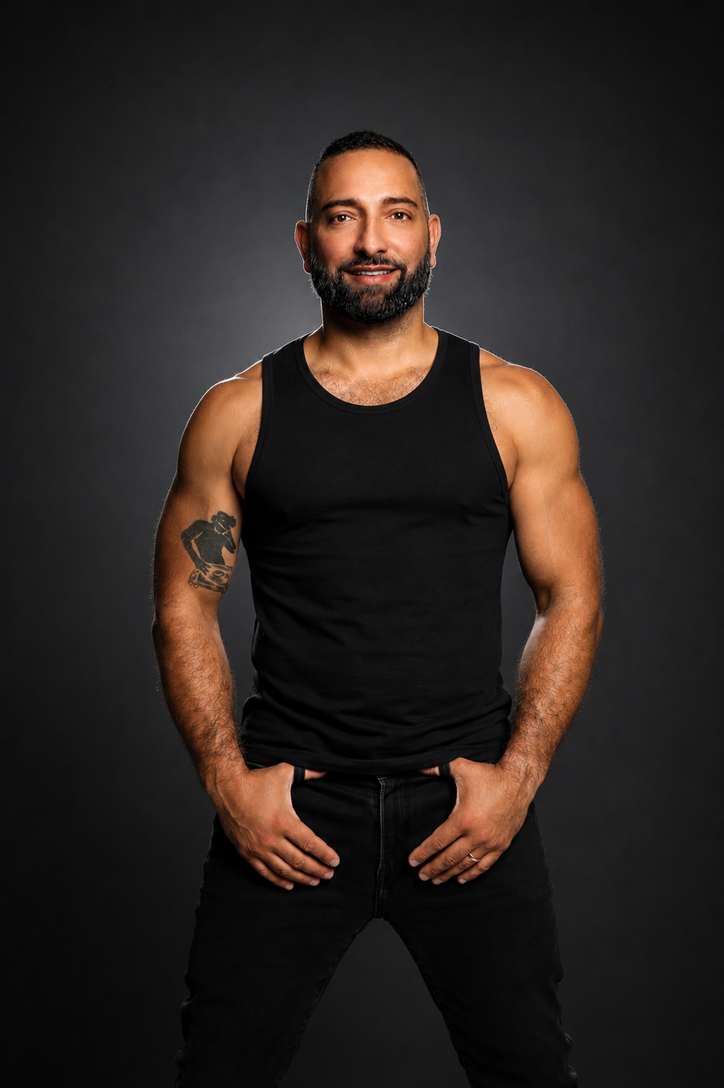 Man with beard and tattoos posing against a dark background, wearing a black sleeveless shirt and black pants.