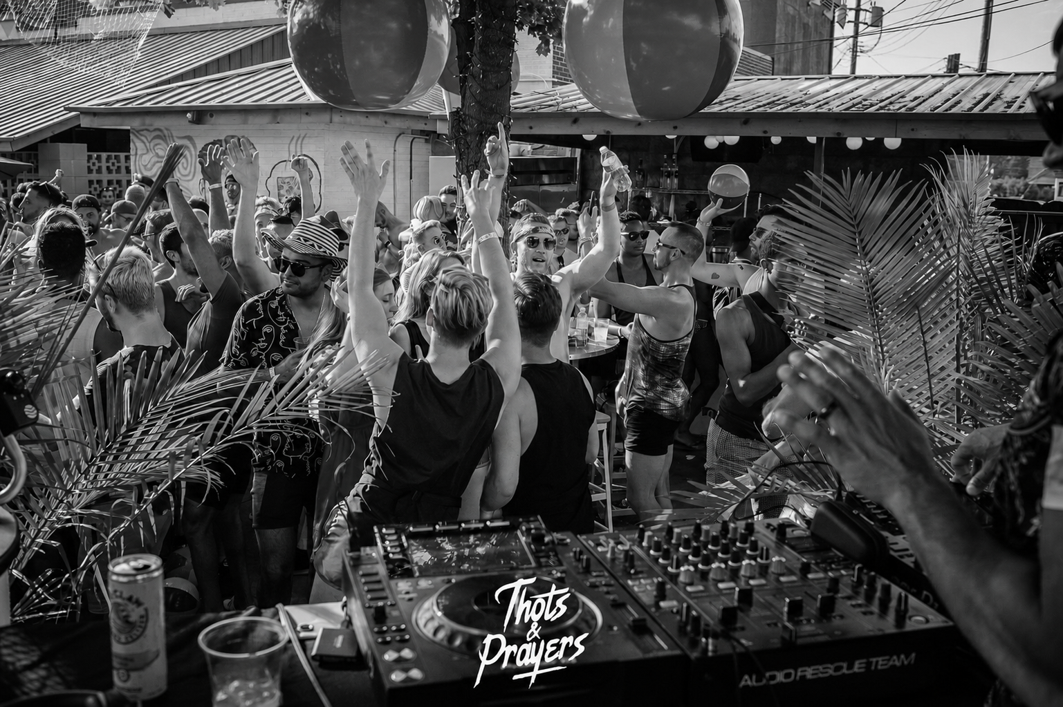 A lively outdoor party at a bar or club, with a crowd of people dancing and enjoying music. A DJ is visible in the foreground, and the scene is decorated with tropical plants and large hanging globes. The photo is in black and white.