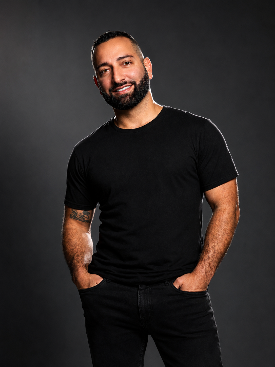 A smiling man with a beard and tattoos, wearing a black t-shirt and black jeans, posing against a dark gray background.