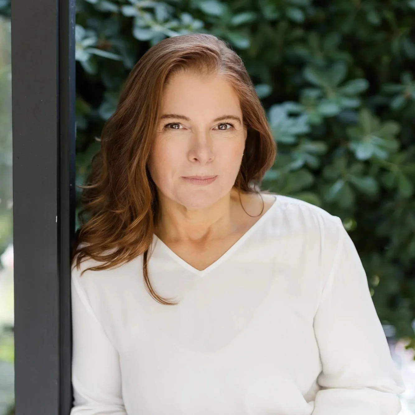 A woman with long, wavy brown hair in a white shirt sitting outdoors near greenery.