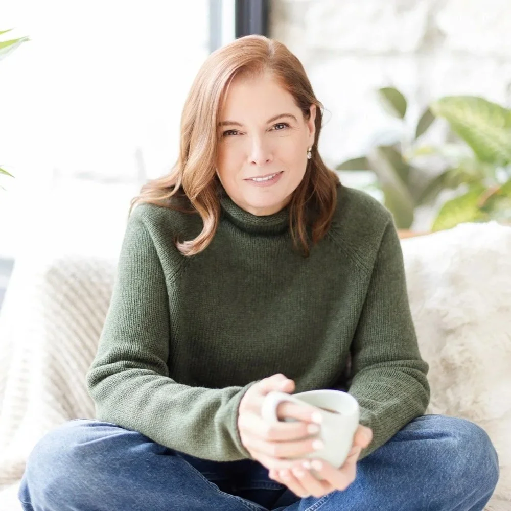 Woman sitting on a sofa holding a white mug, smiling and squinting indoors with green plants and light coming through windows in the background.