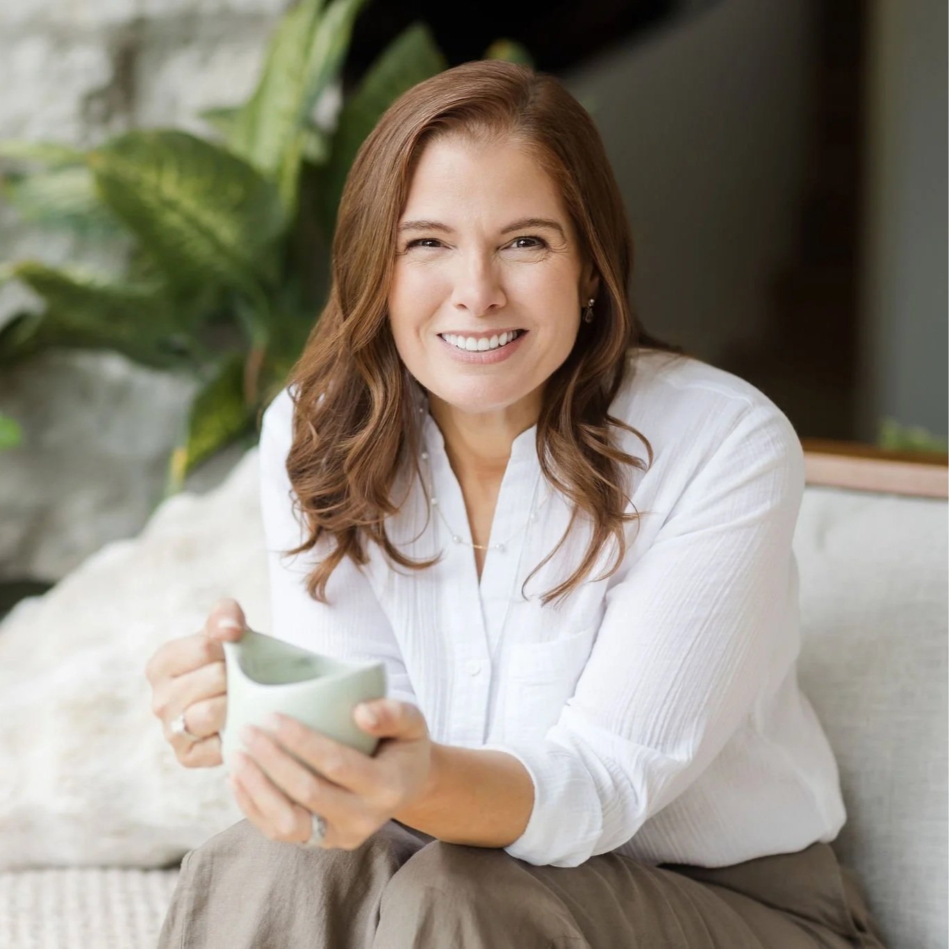 A woman with light skin and shoulder-length brown hair smiling while holding a white mug and sitting on a couch in a room with green plants.