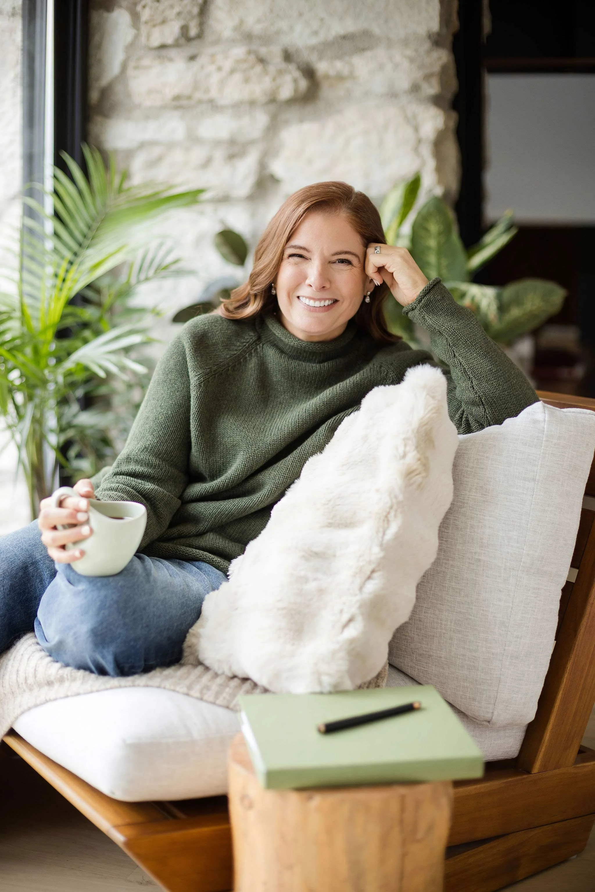 A woman with reddish hair smiling and winking, sitting on a cozy chair, holding a cup of coffee, surrounded by indoor plants and a rustic stone wall background.