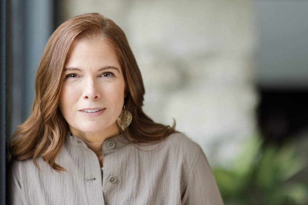 A woman with shoulder-length brown hair and earrings, wearing a beige shirt, smiling and standing indoors near a window.