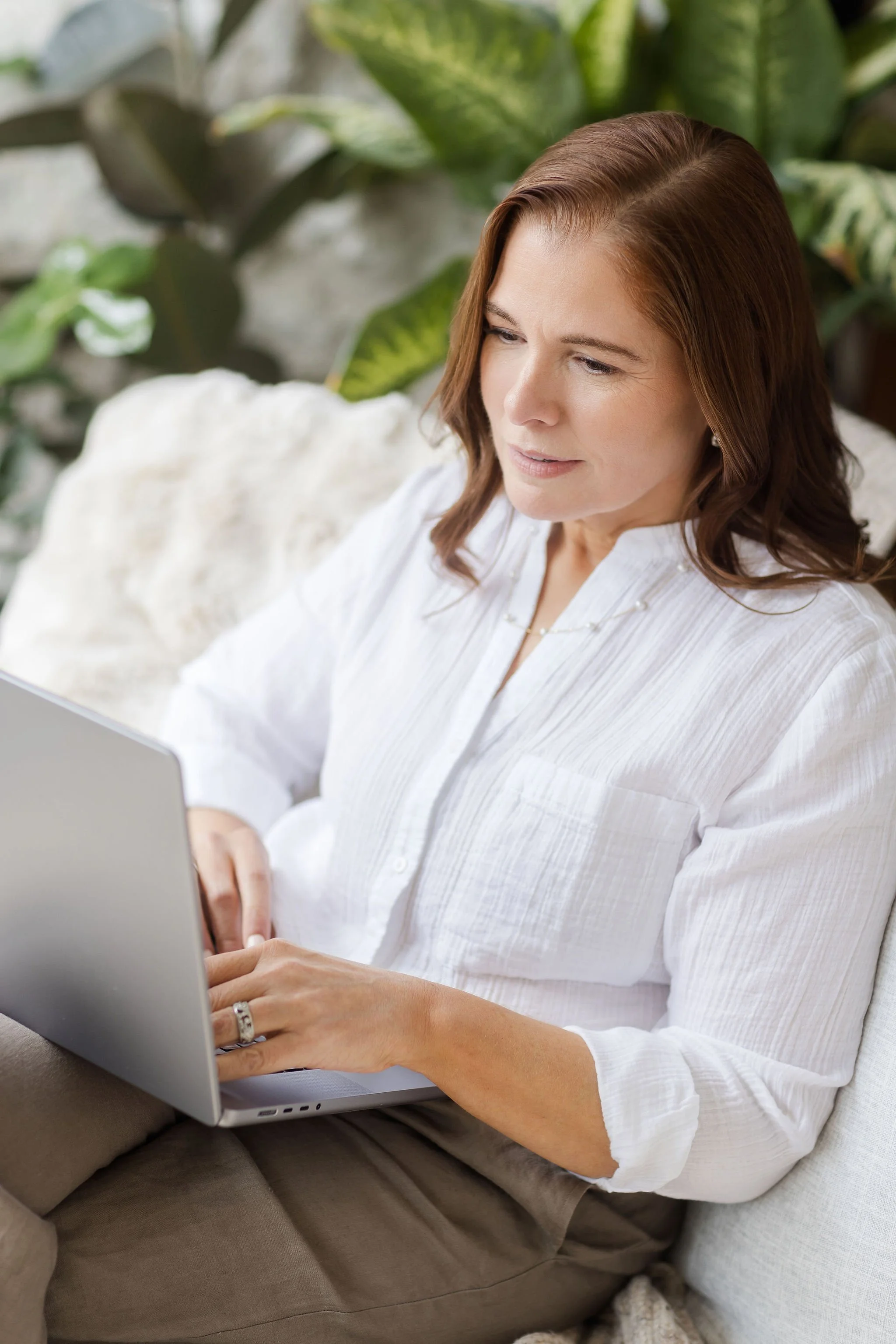 A woman with brown hair and wearing a white blouse sitting on a couch using a silver laptop, with large green houseplants behind her.