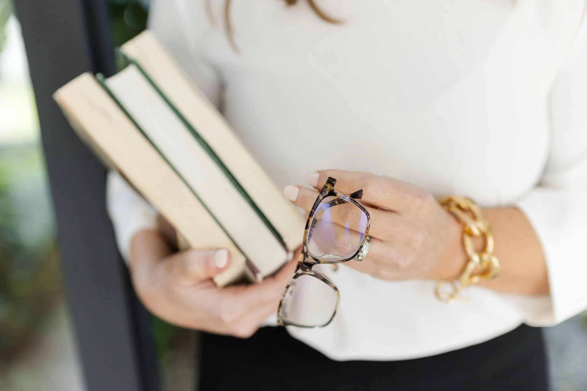 A person holding a stack of books with a pair of glasses in their hand, wearing a white shirt and gold jewelry.