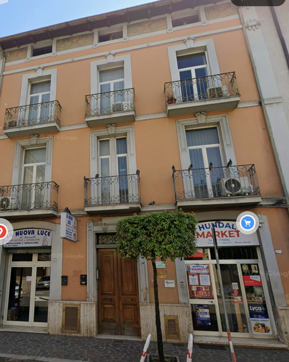 Edificio con tre piani, finestre con balconi in ferro battuto, negozio al pianterreno chiamato 'HUNDAL MARKET' e 'NUOVA LUCE'.