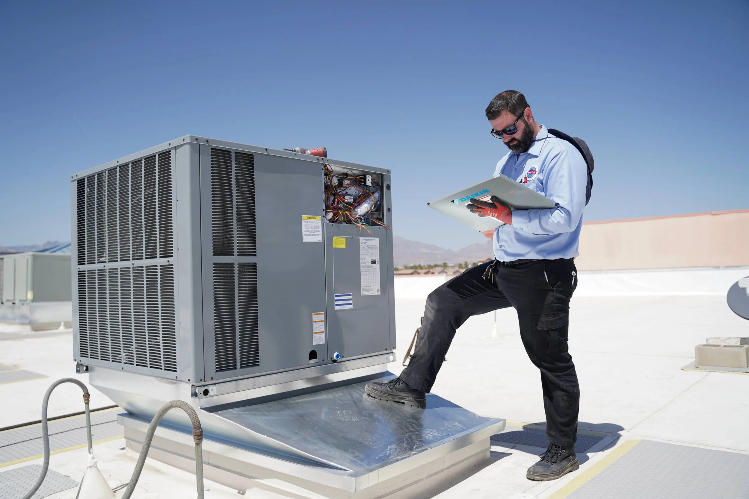 Technician inspecting and working on an air conditioning unit on a rooftop, with a clear blue sky and distant mountains in the background.