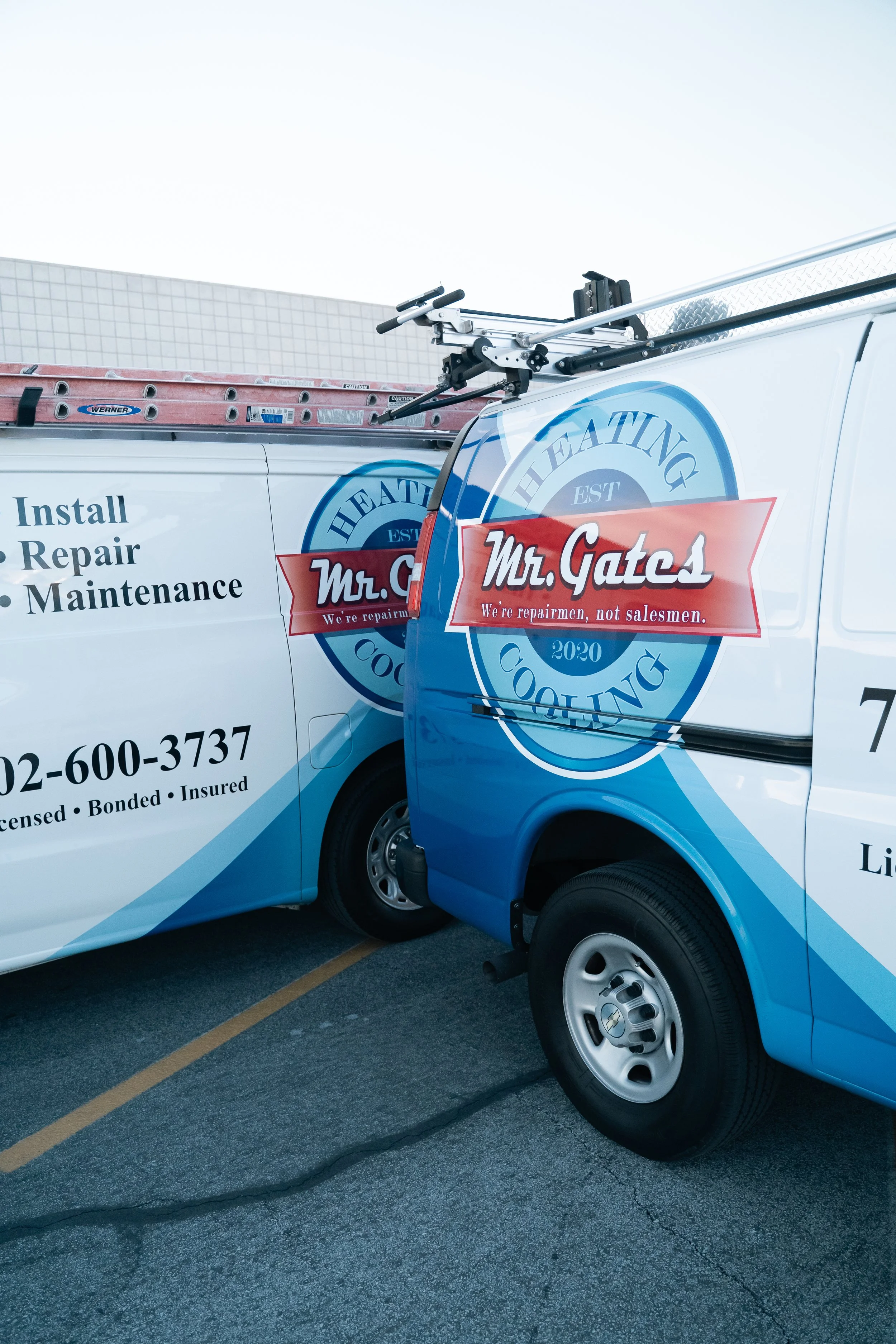 Two repair service trucks labeled Mr. Gates are parked side by side, with ladders on top, at a parking lot. The trucks display contact information and services offered such as install, repair, and maintenance.
