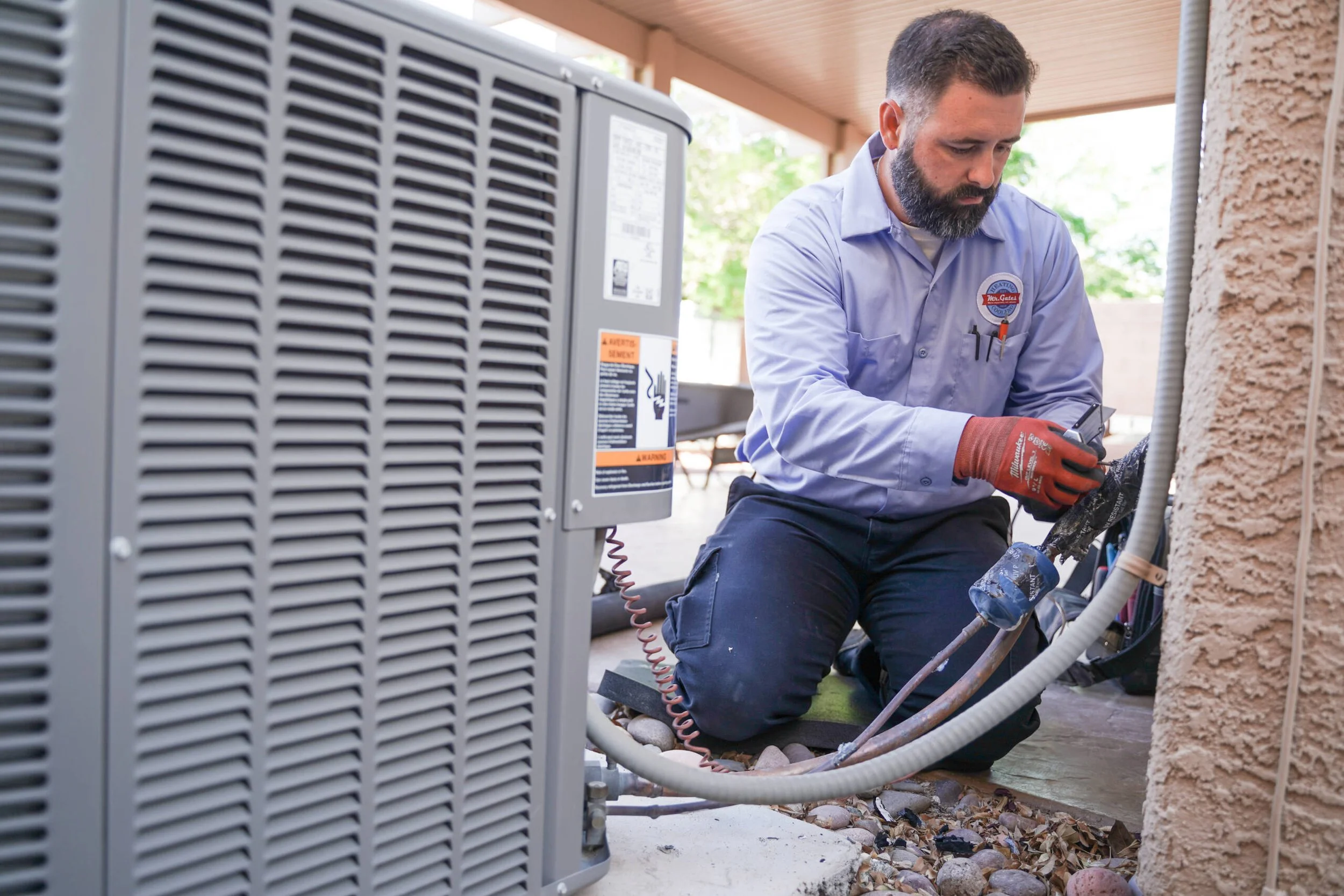 A technician wearing a blue shirt, red gloves, and kneeling on the ground working on outdoor electrical or HVAC equipment near a beige stucco wall in a residential area.