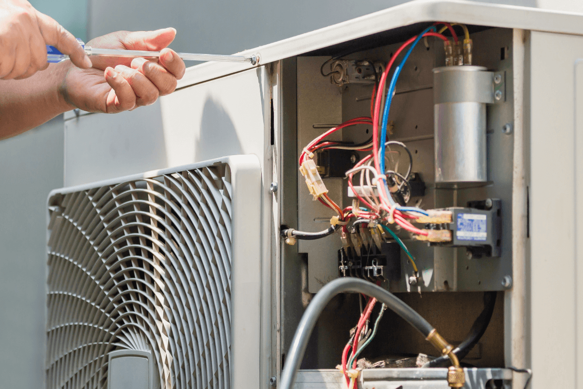 Person repairing or inspecting the internal components of an air conditioning unit, with a screwdriver in hand and exposed wiring visible.