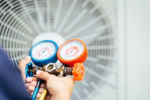 Person holding a manifold gauge set with blue and orange gauges in front of an air conditioning condenser coil.