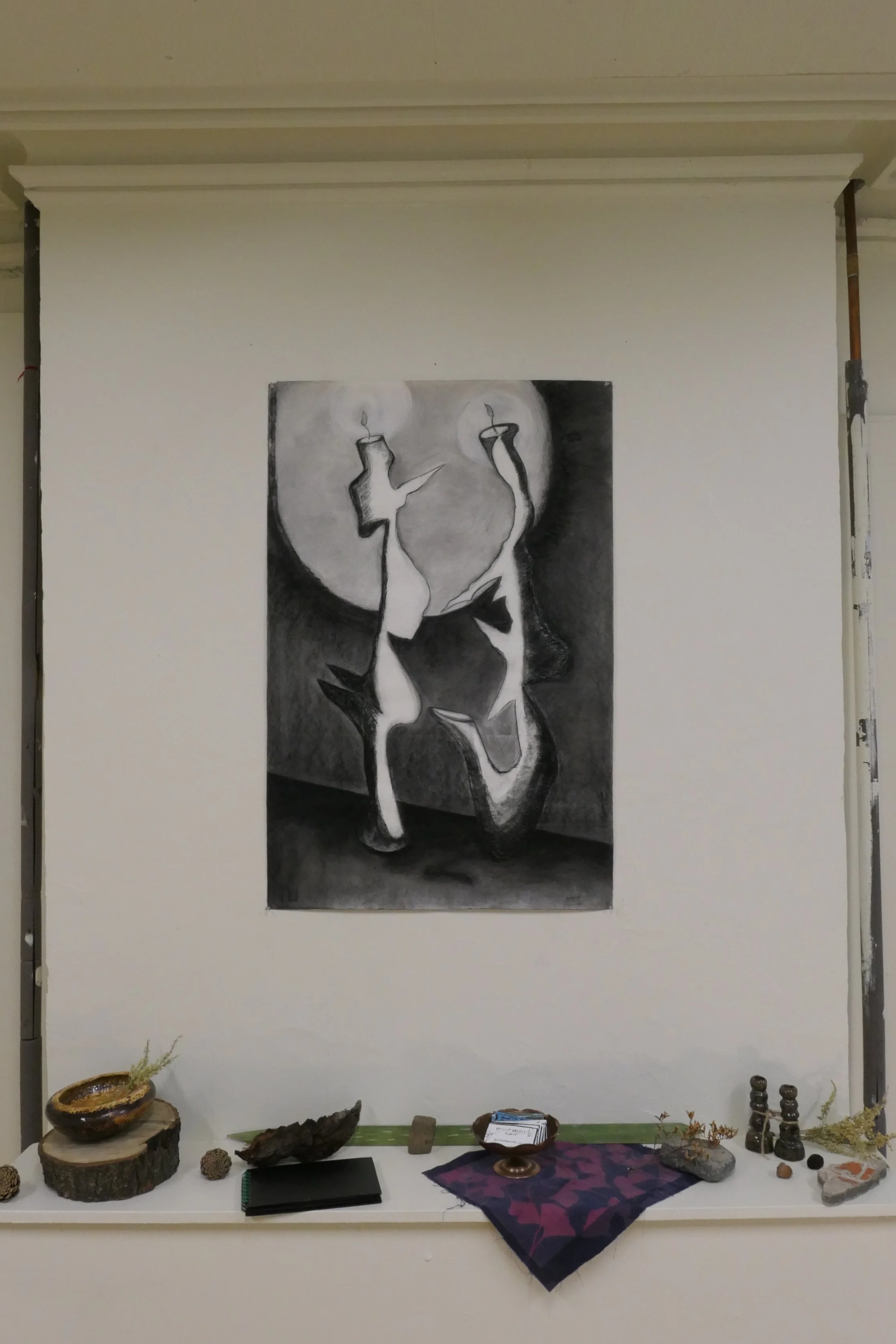 An art display with a black and white abstract drawing of two candles on a white wall. Below, a table showcases wooden bowls, stones, small sculptures, plants, and a red cloth.