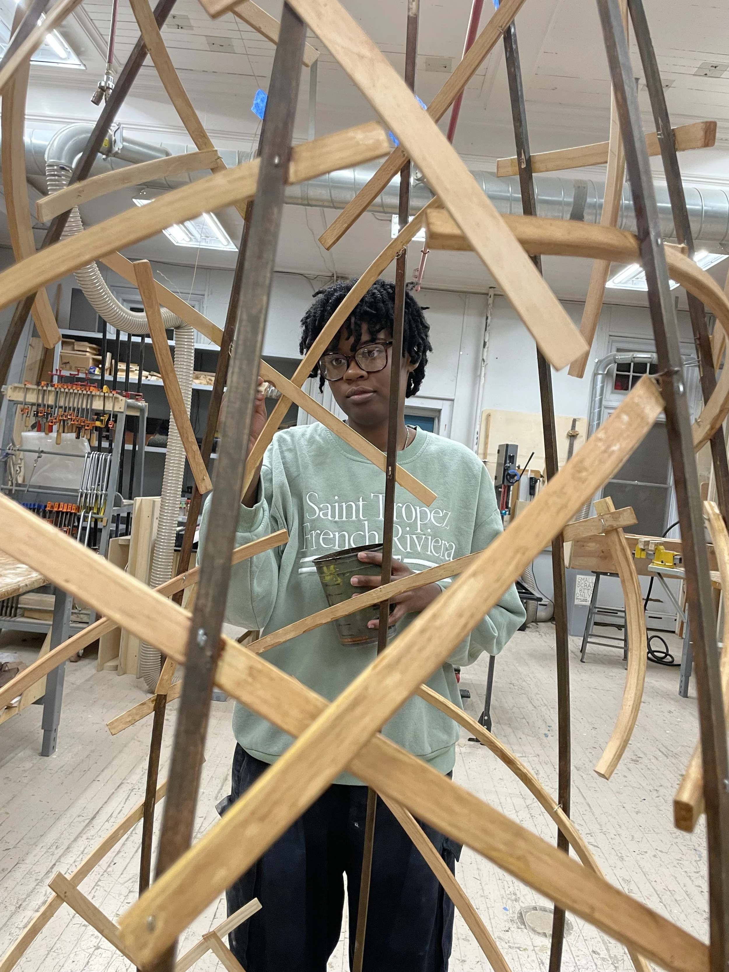 An artist with glasses working on a large wooden art piece in a workshop. The artist is wearing a light green sweatshirt with text on it. The workshop has tools, shelves, and ductwork visible in the background.
