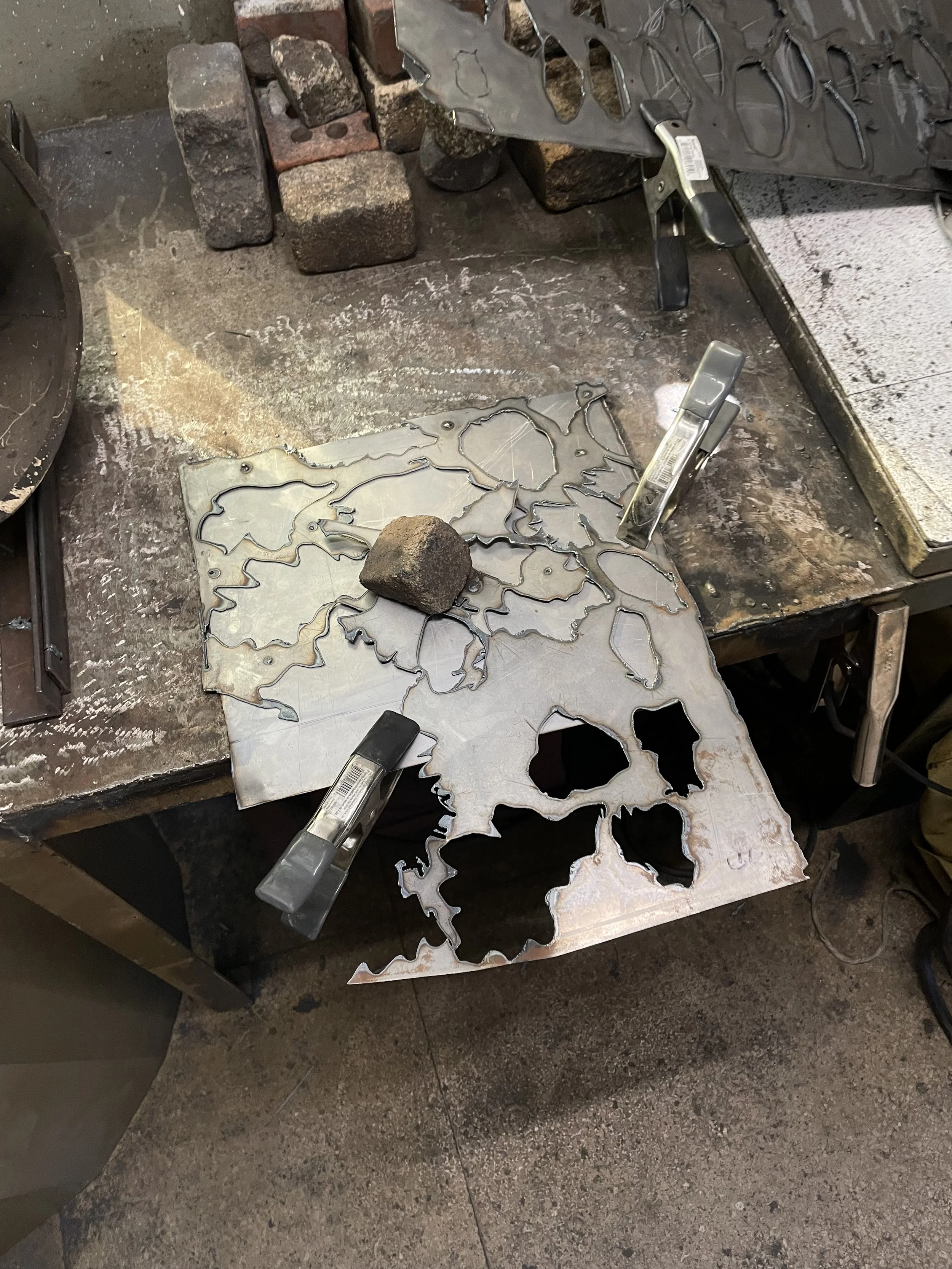 Work surface with cutout metal sheets, a brick and rock, and tools with black handles. The surface is stained and slightly rusty.