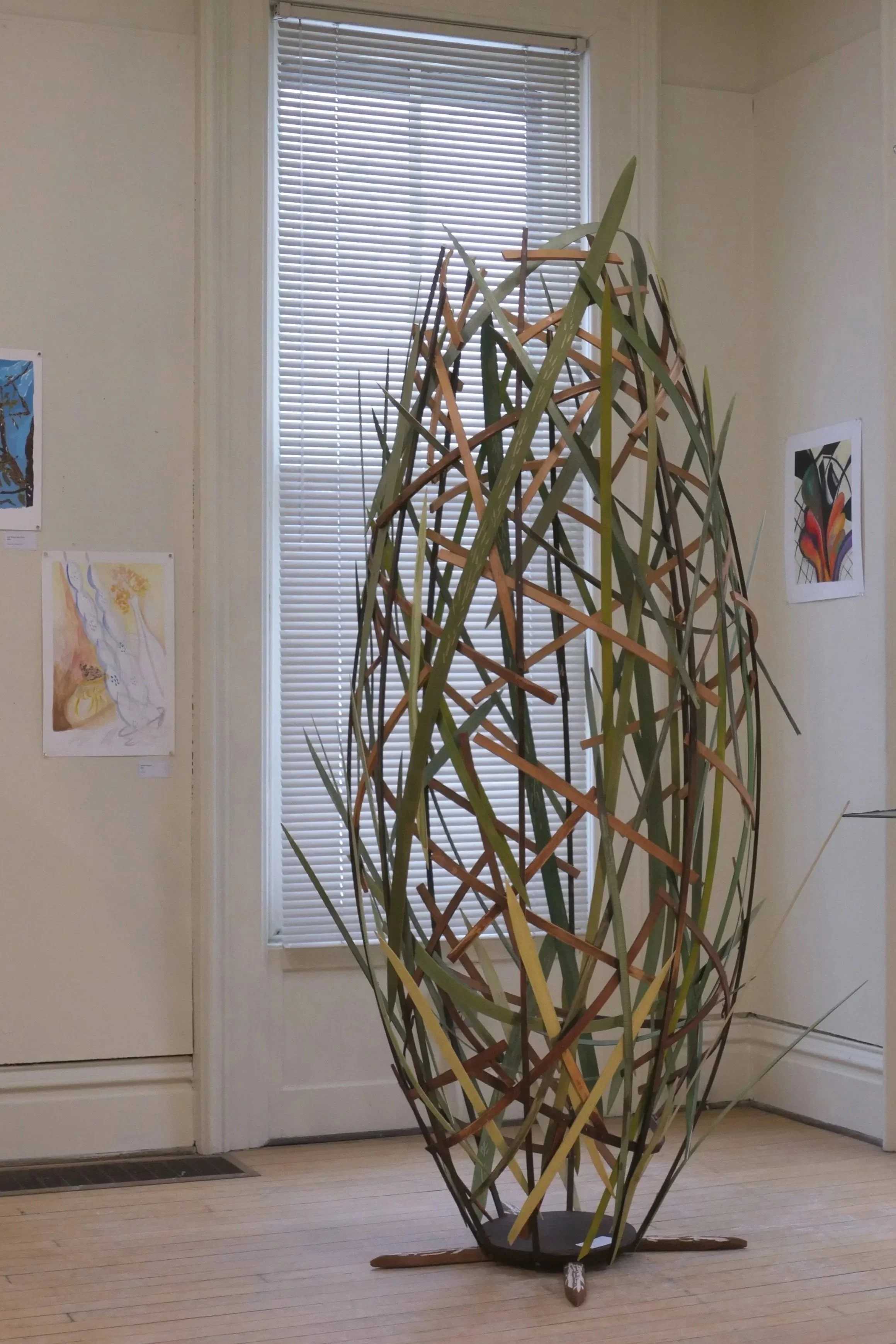 A large abstract sculpture made of woven wooden strips is displayed indoors near a window with blinds. The sculpture is set on a wooden floor with a white wall in the background, which features framed artwork.
