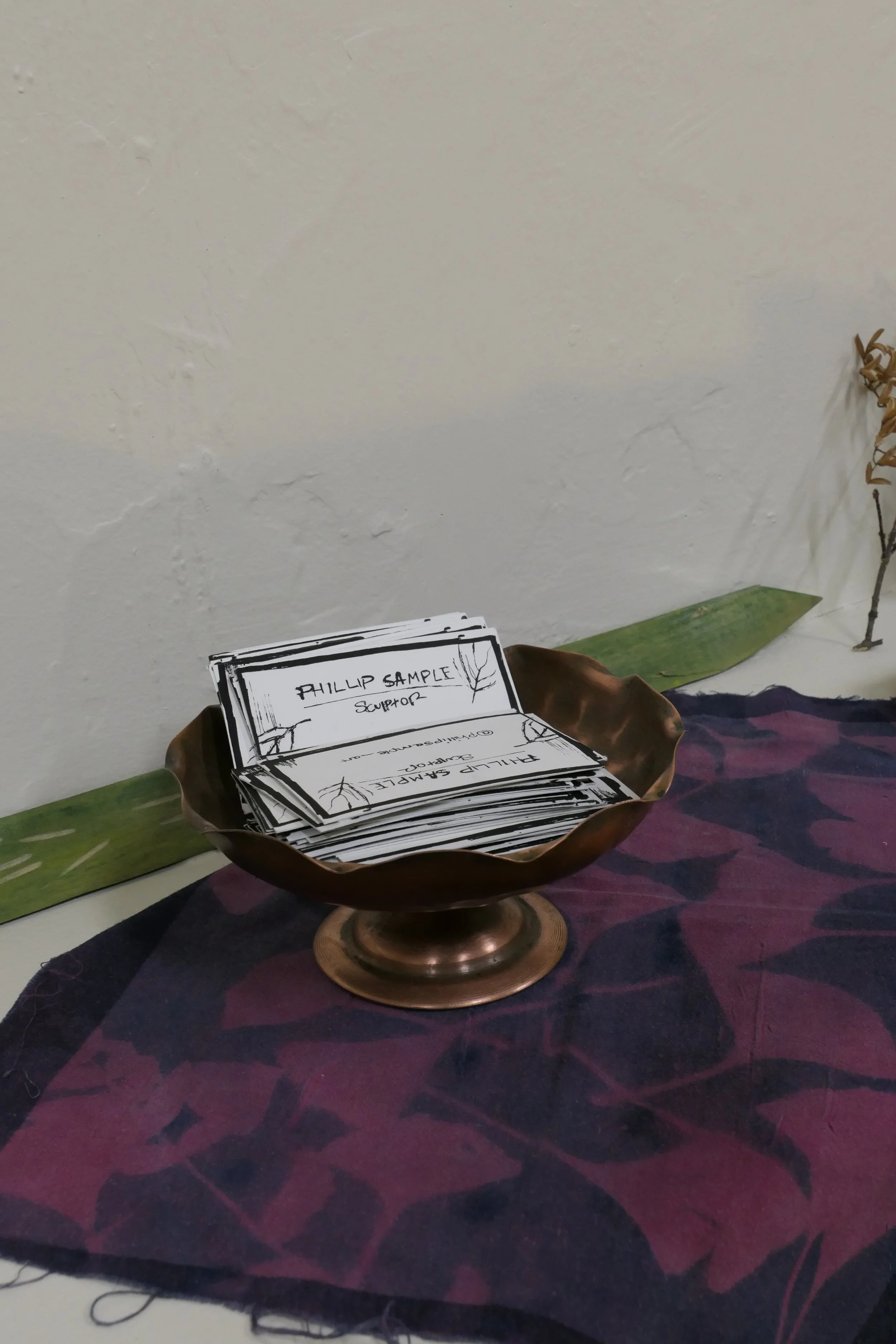 A  bowl filled with black and white printed business cards, labeled with 'Phillip Sample' rests on a dark purple and red patterned cloth, with a large green leaf and a dried flower arranged nearby against a plain white wall.