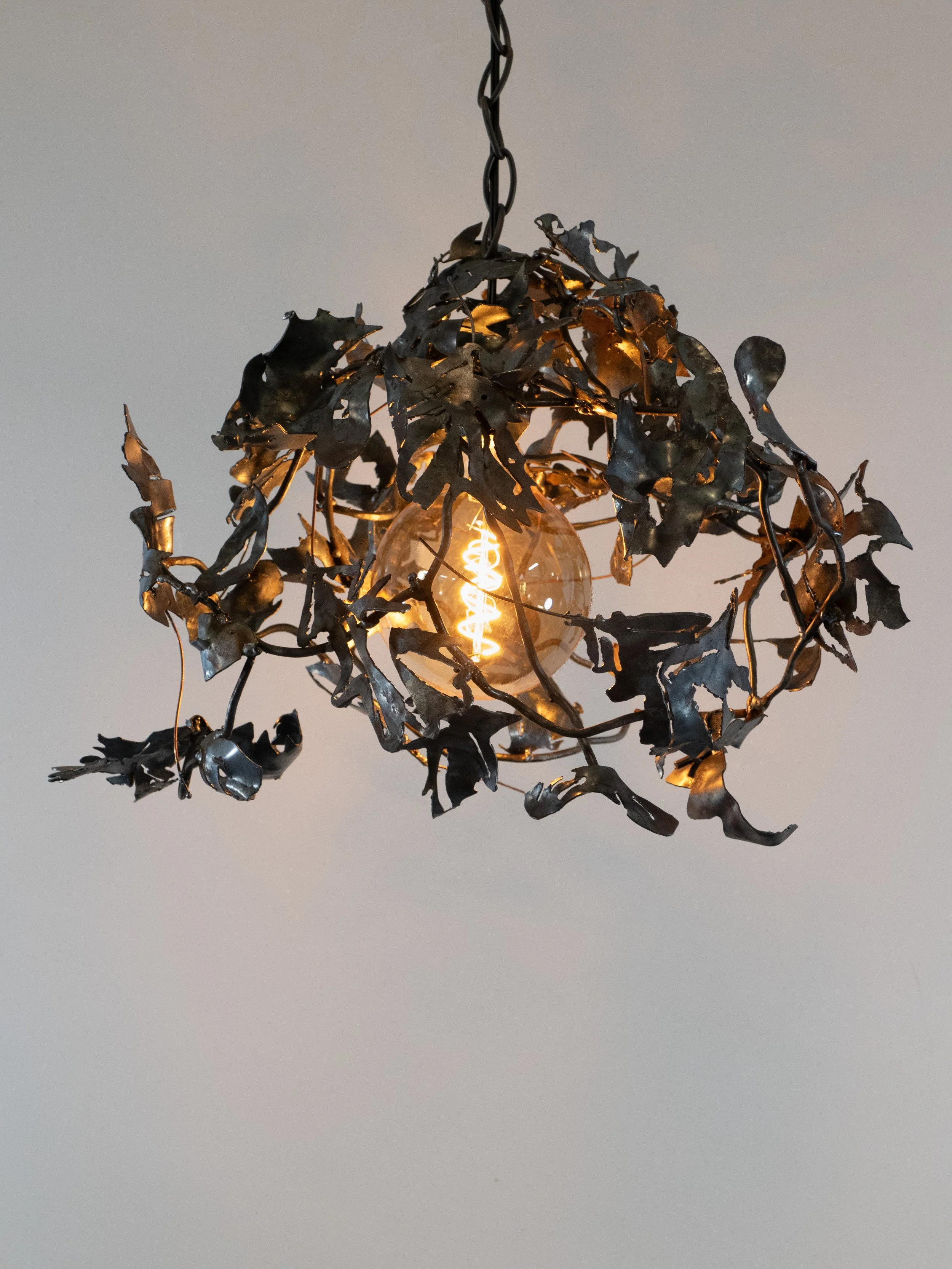 A hanging light fixture with a decorative metal frame resembling leaves surrounding an exposed vintage-style filament bulb.