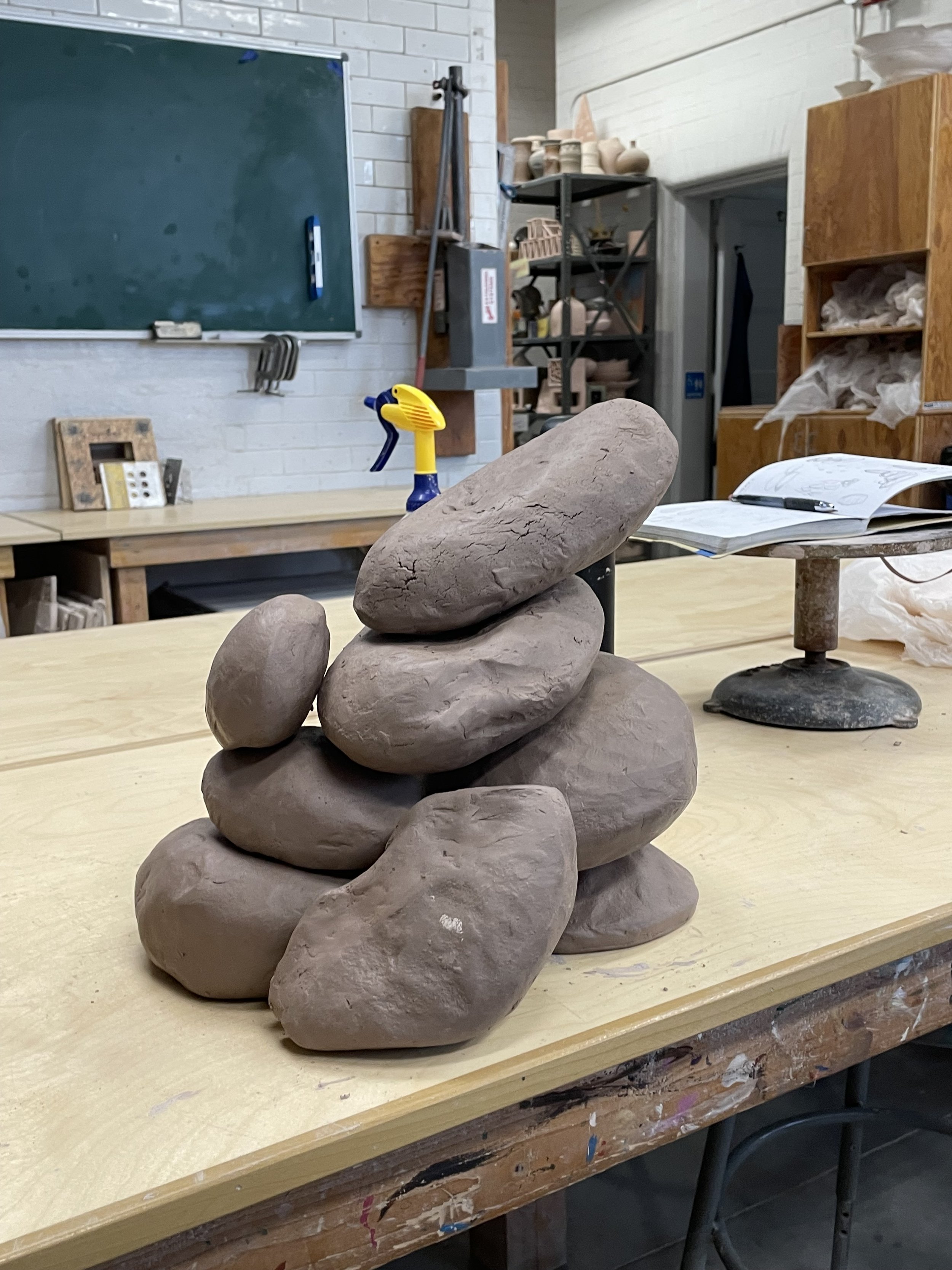 Ceramic stones stacked on a wooden table in an art studio, with a workspace background including a chalkboard, shelves, and art supplies.