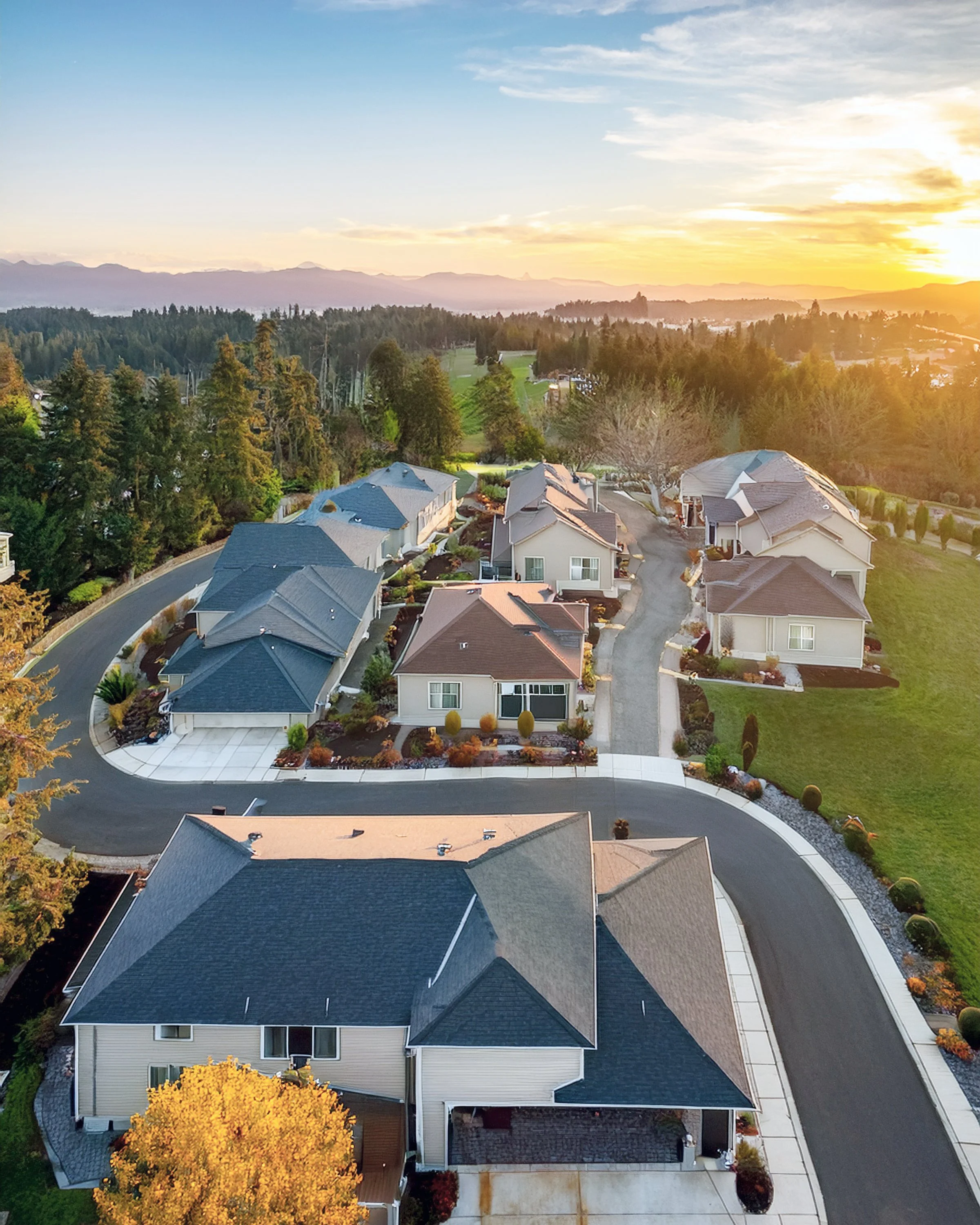 An aerial view of a suburban neighborhood during sunset showing houses with dark roofs, a curved road, and trees with autumn foliage, mountains in the background.