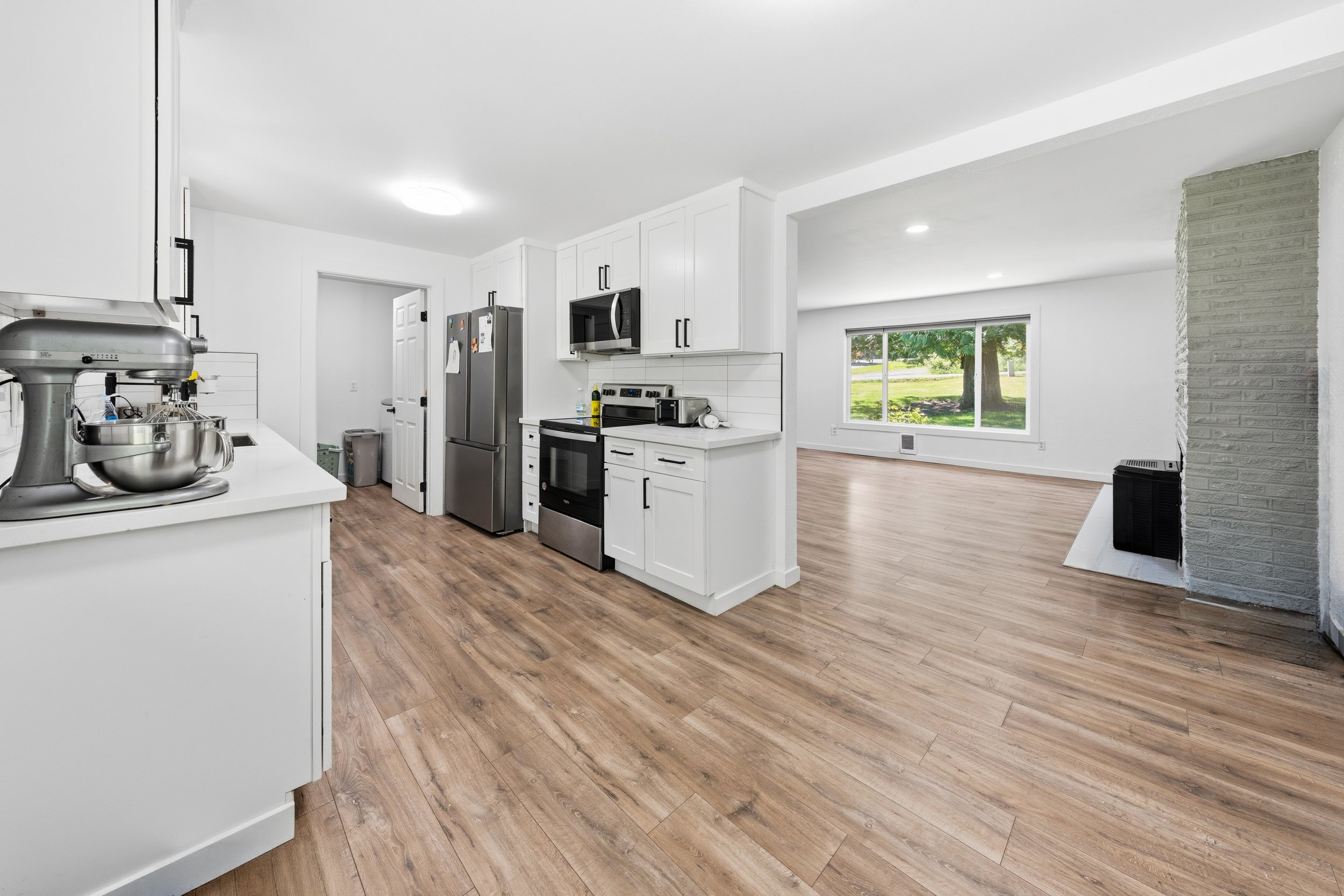 Modern kitchen with white cabinets, stainless steel appliances, and wood flooring, open to a bright living area with large window showing a green outdoor scene.