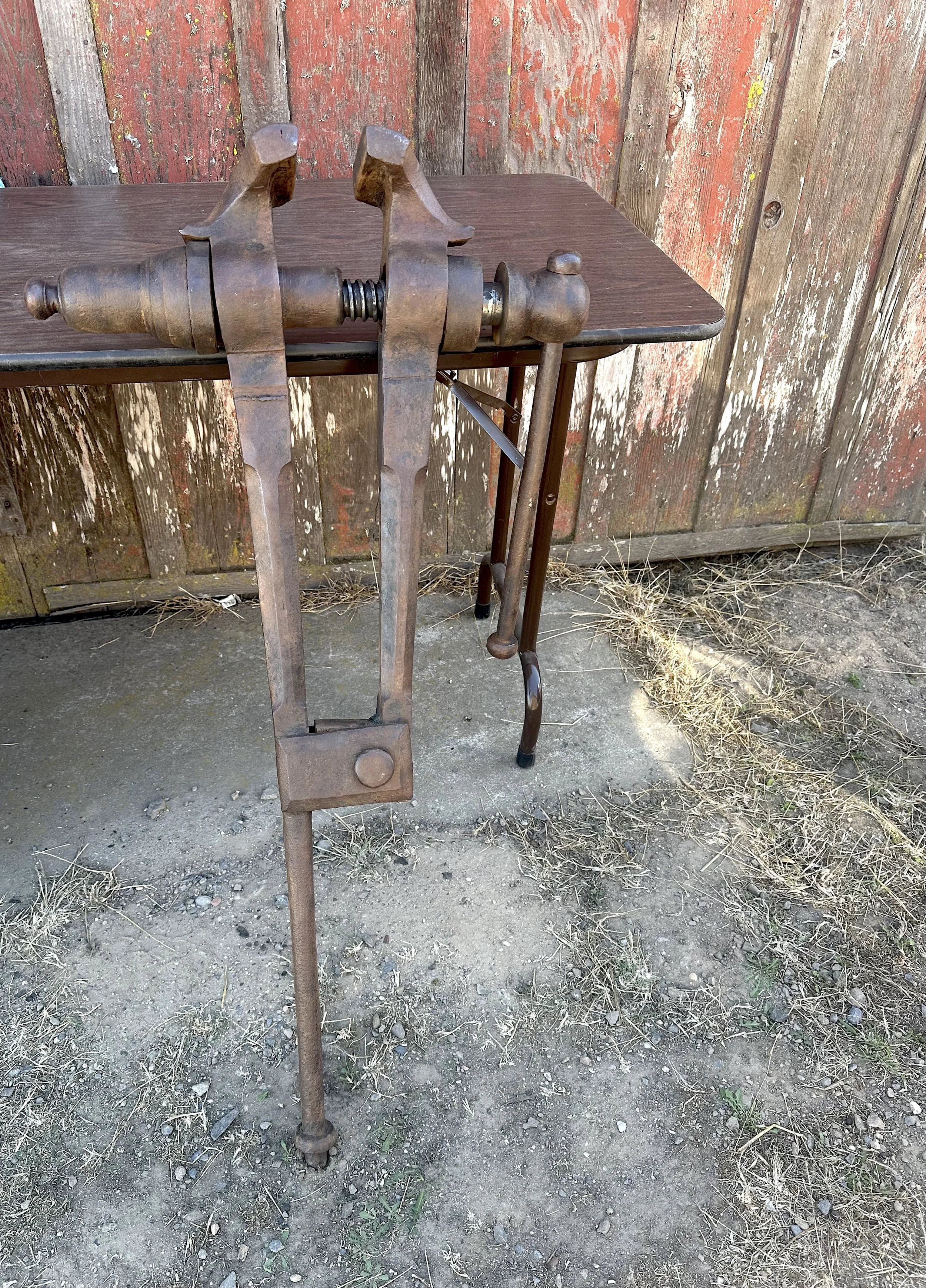 An old, metal vise with a post leg leaning against a table.