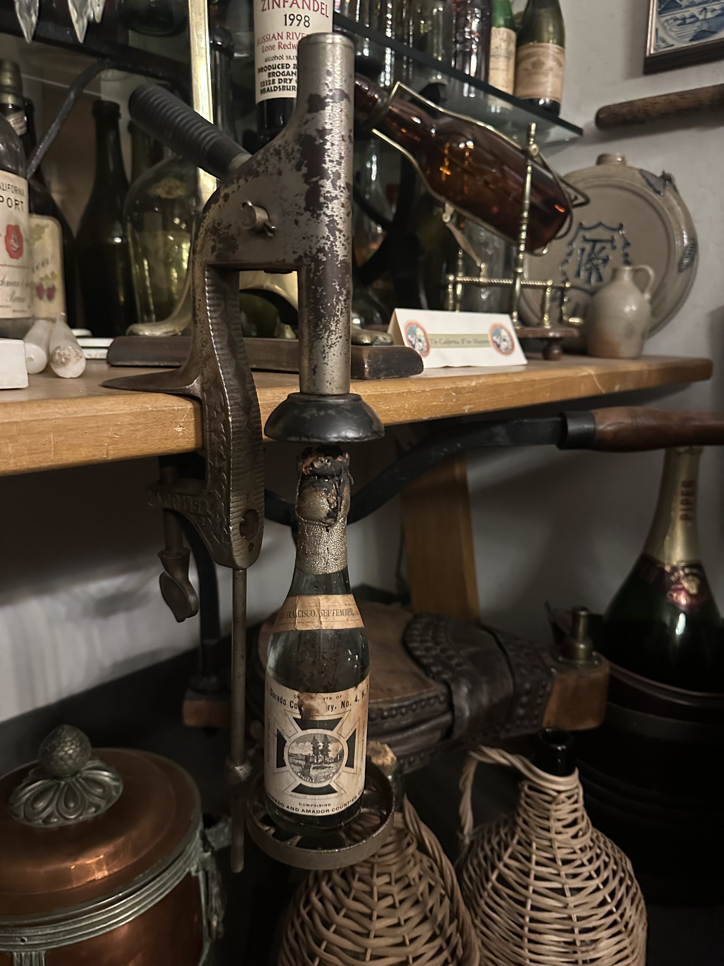 A metal bottle capper atop an old bottle. It is attached to a shelf.