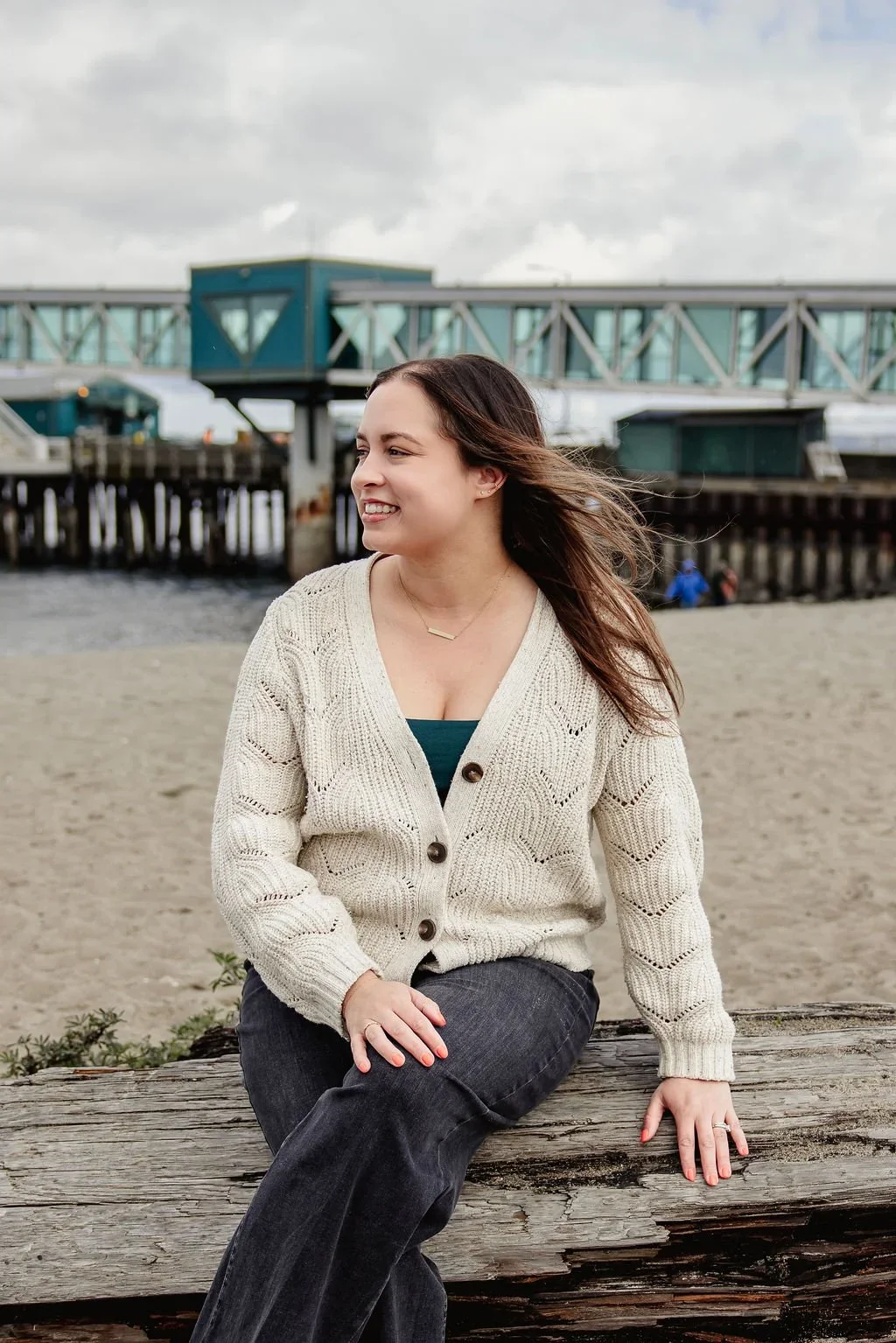 A therapist for moms sitting on a wooden log at the beach, smiling and looking to her left. She is wearing a cream-colored cardigan over a green top, with dark jeans. In the background, there is a pier with a bridge and overcast sky.