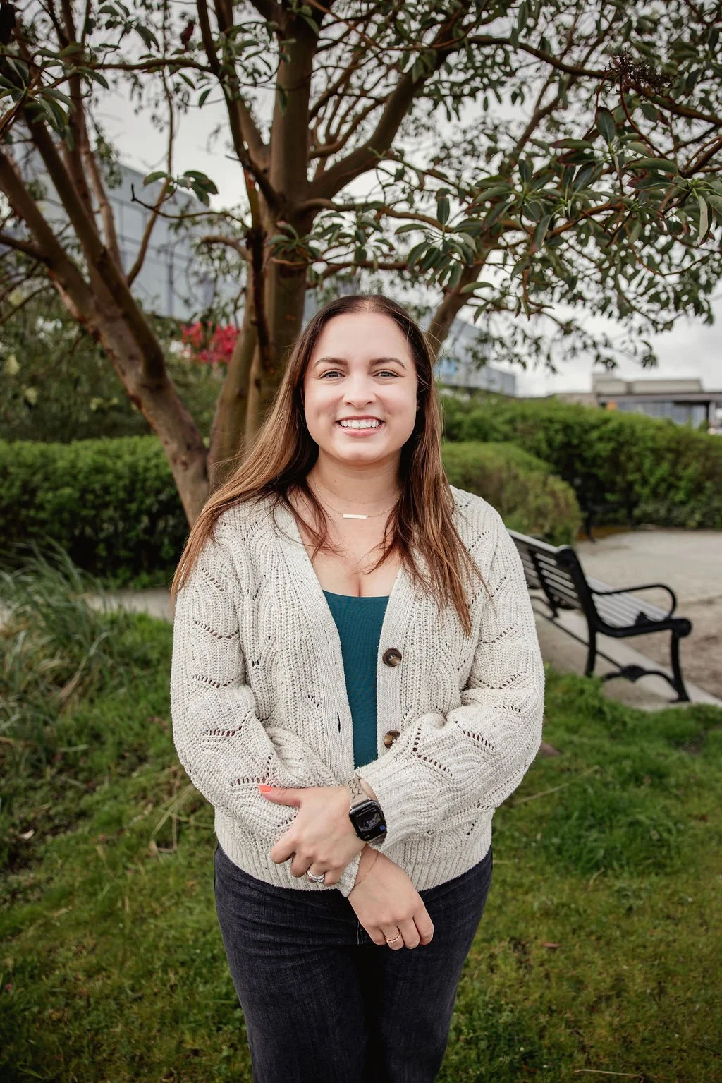 A smiling therapist with long brown hair standing outside in Edmonds in front of a tree, with benches and greenery in the background.